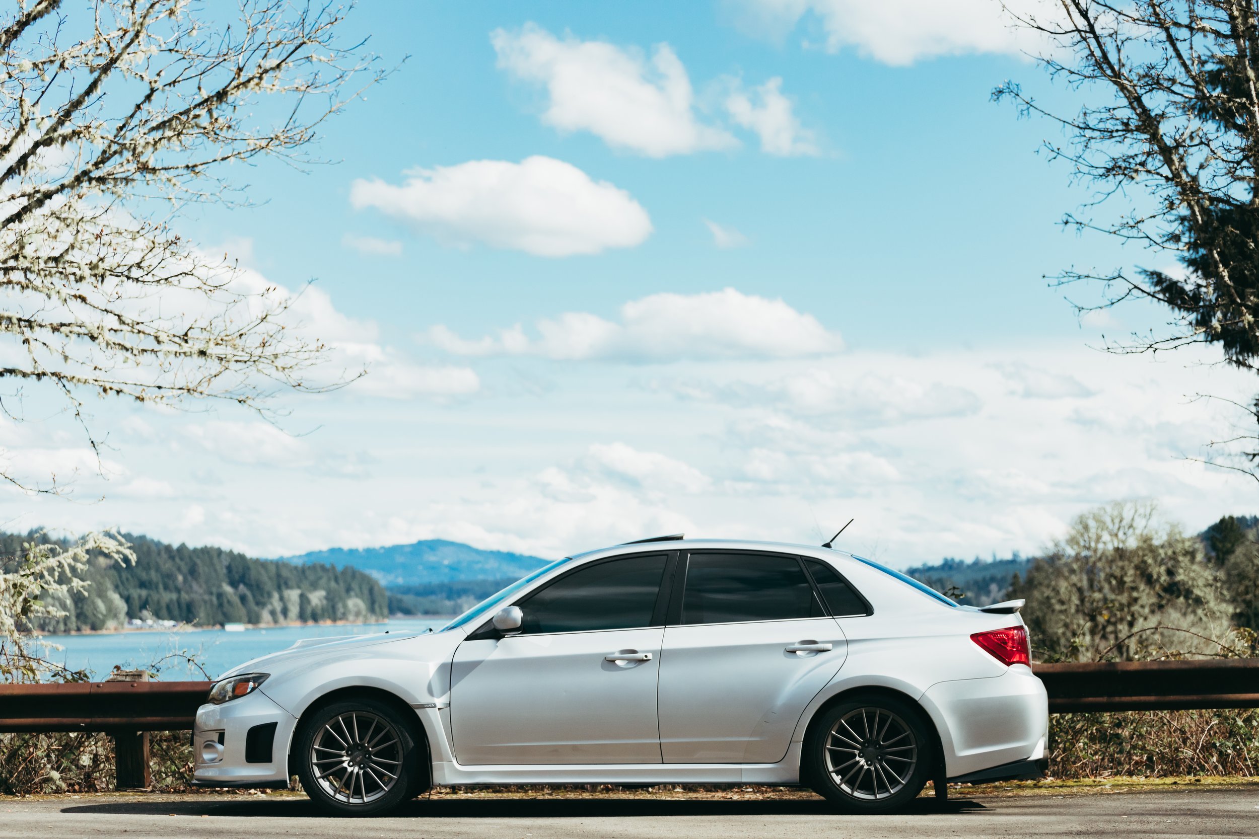 A silver sedan parked beside a guardrail on a scenic roadside with a lake and forested mountains in the background under a partly cloudy sky.