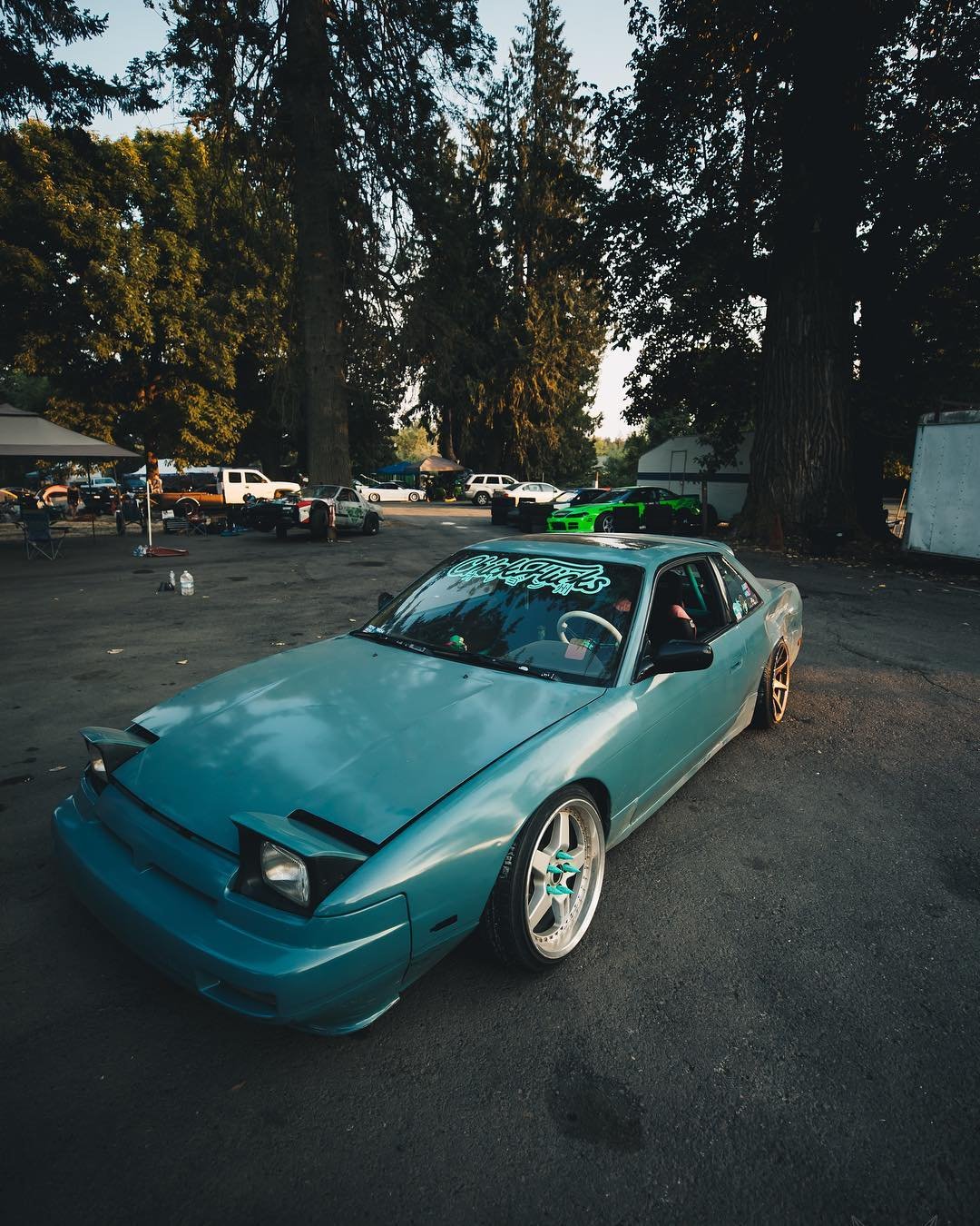 A light blue, modified sports car with pop-up headlights parked in a lot surrounded by trees and other vehicles, some of which are high-performance cars.