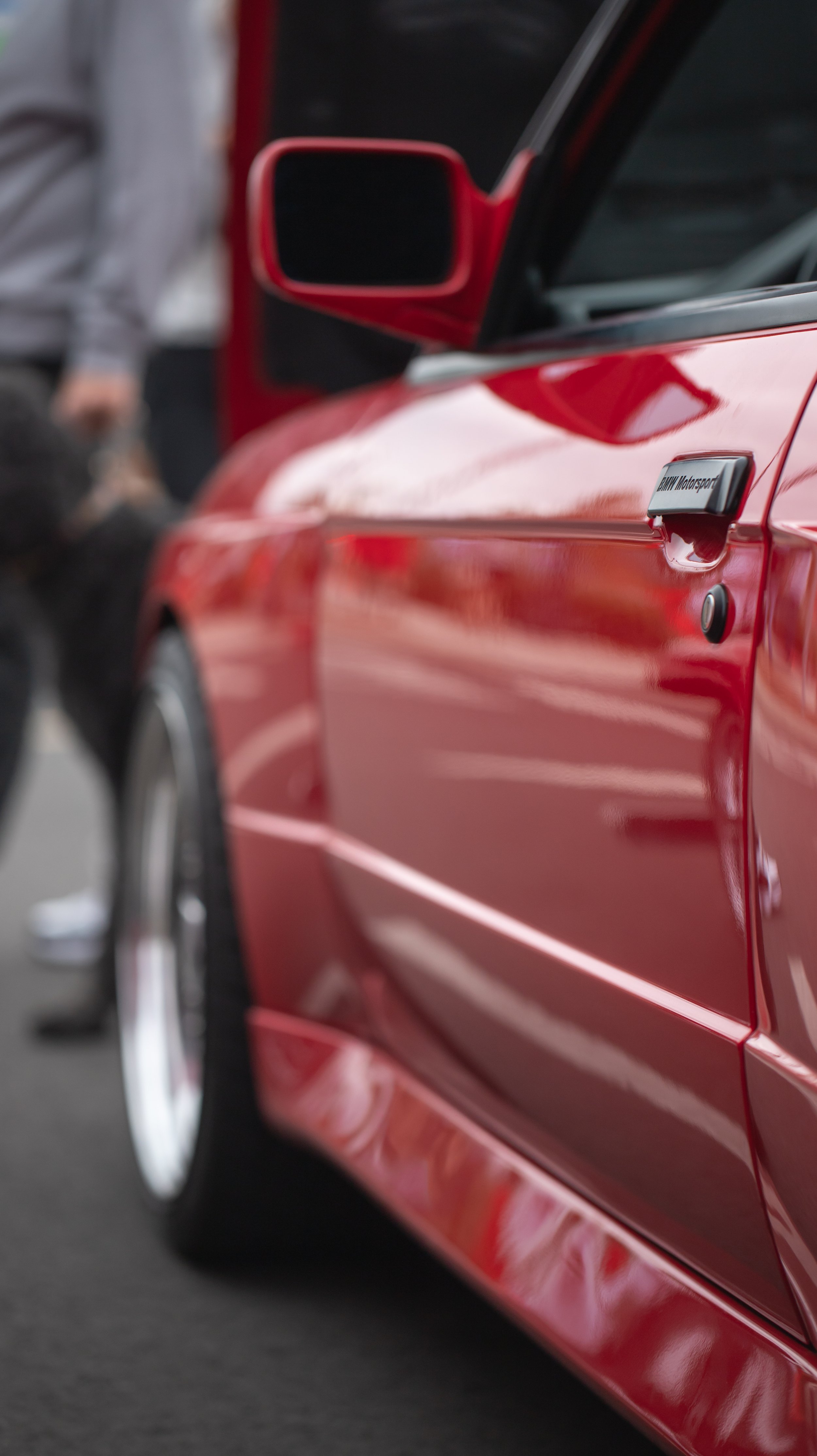 Close-up of a red sports car with a 'BMW Motorsport' emblem on the door, reflecting light and showing a blurred person in the background