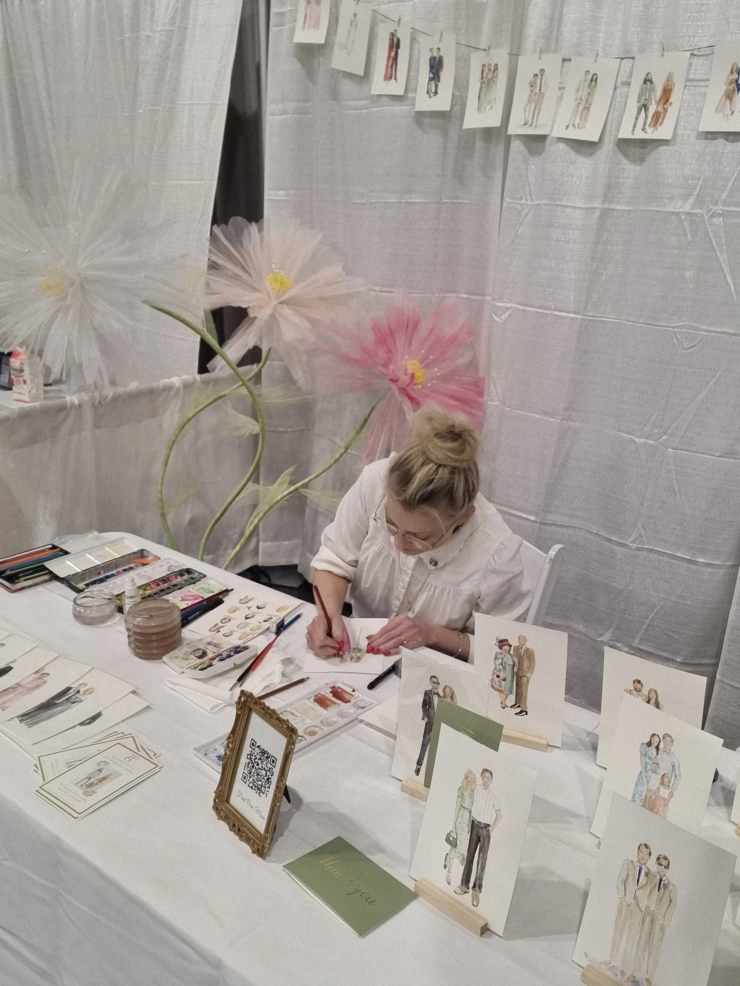 woman dressed in white sitting at a table painting watercolor paintings of event and wedding guests for the Engaged Kansas City Bridal Show