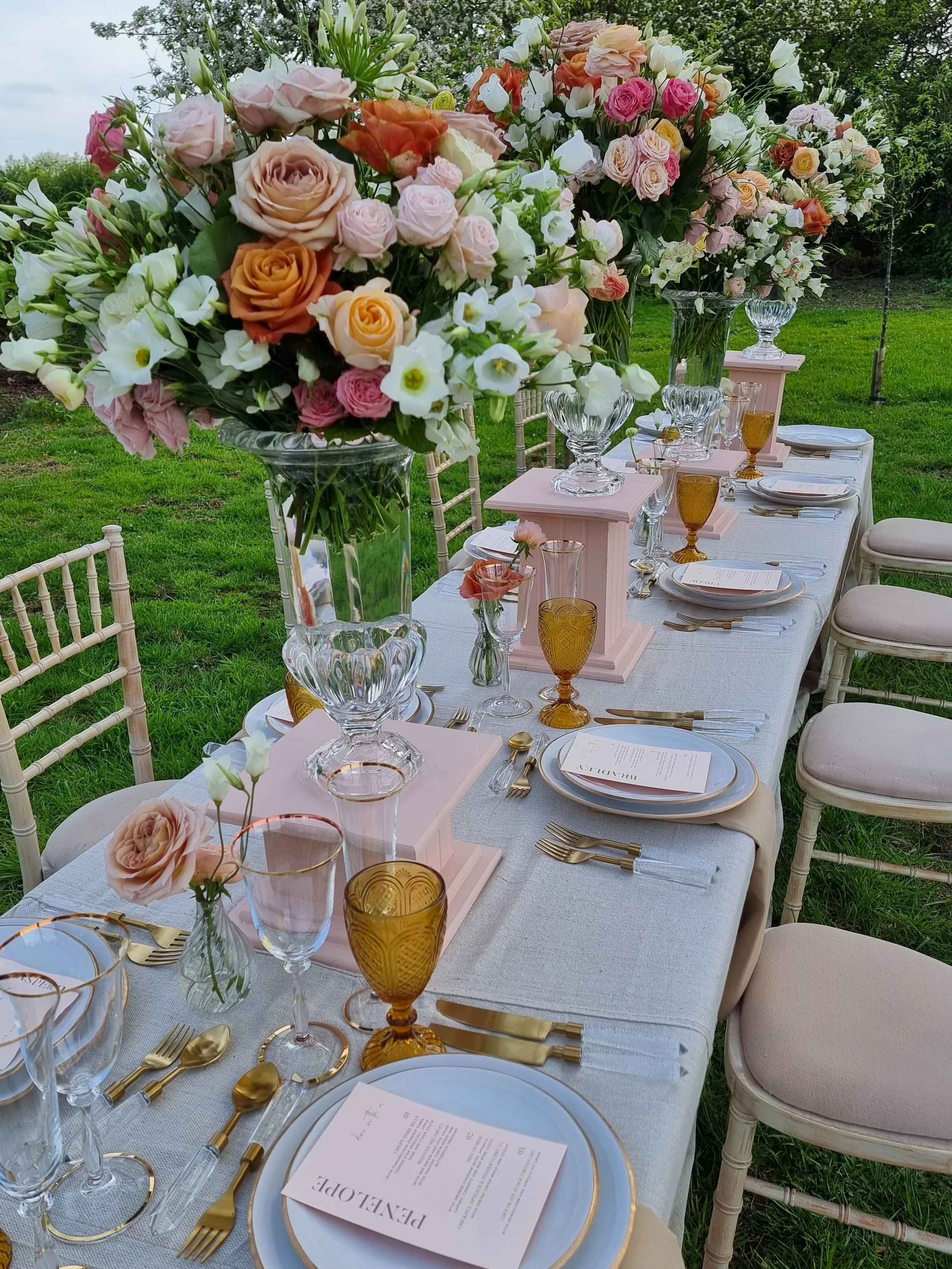 a wedding table set for the reception with large floral arrangements in the middle sitting on plynths and a pink table cloth.
