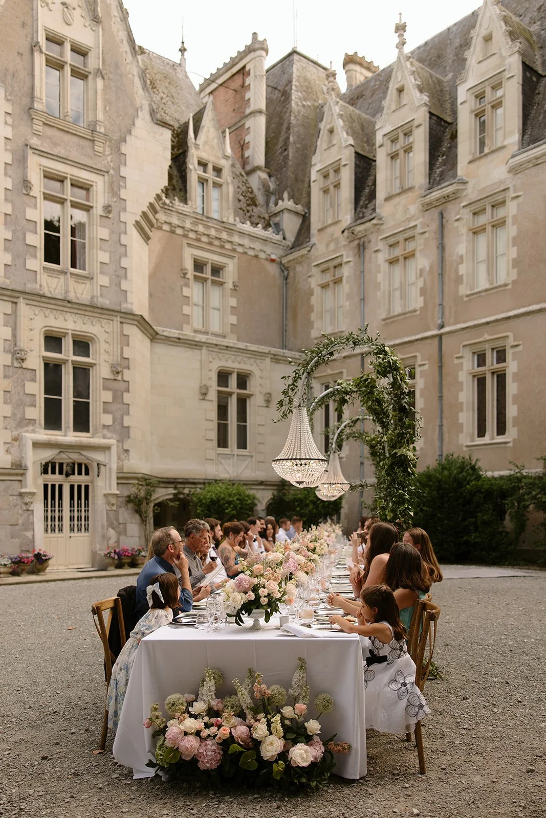 People dining at a Wedding table at Chateau Allure du Lac for a wedding in France in June with hanging chandeliers over the table and flowers.