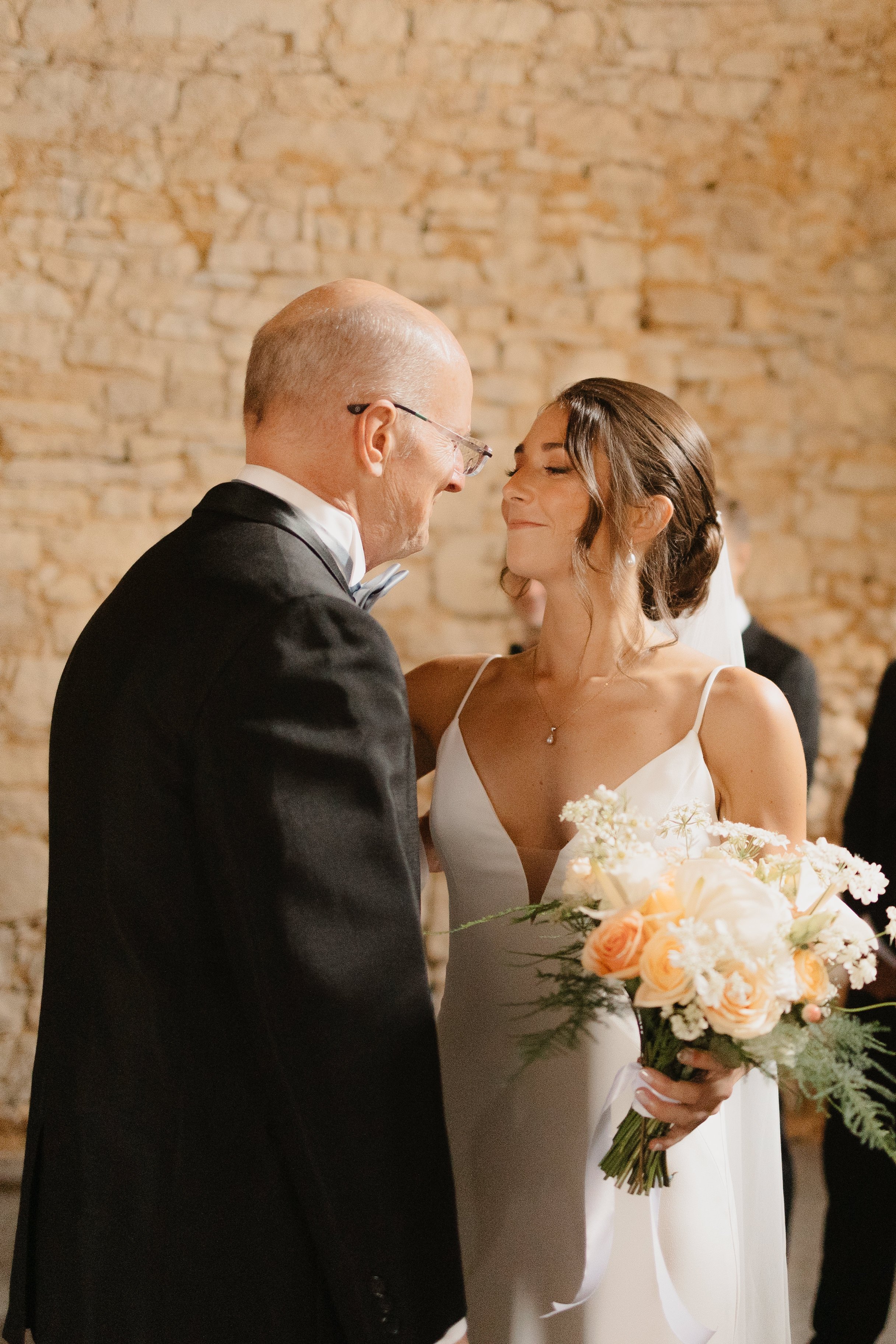 The bride and her father embrace after walking down the aisle as she holds her bouquets of peach and white flowers in a row at a chateau in France for her destination wedding in France