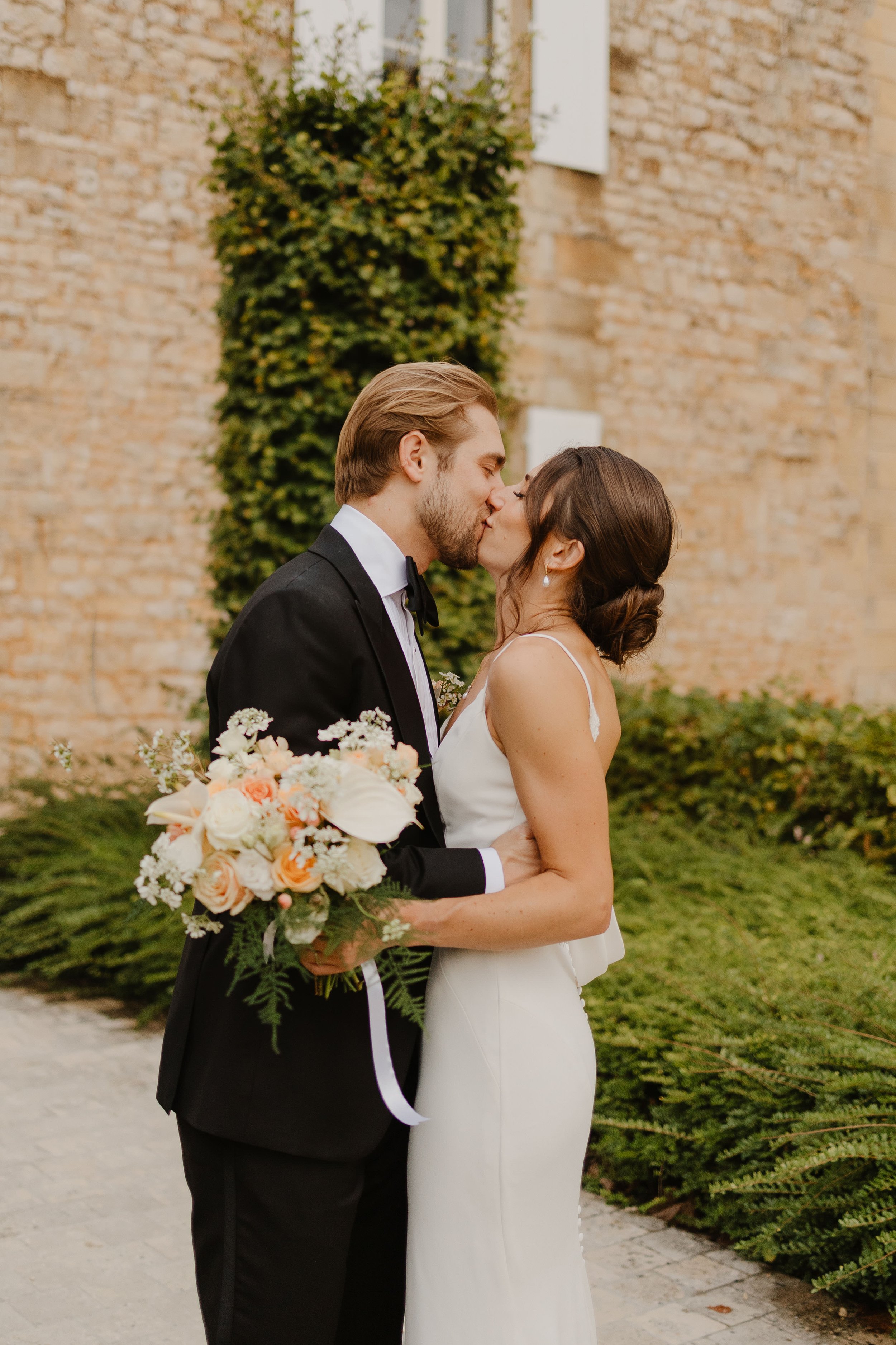 The bride and groom stand in front of the Chateau kissing with her floral bouquet of flowers of peach and white flowers in a row at a chateau in France for her destination wedding in France