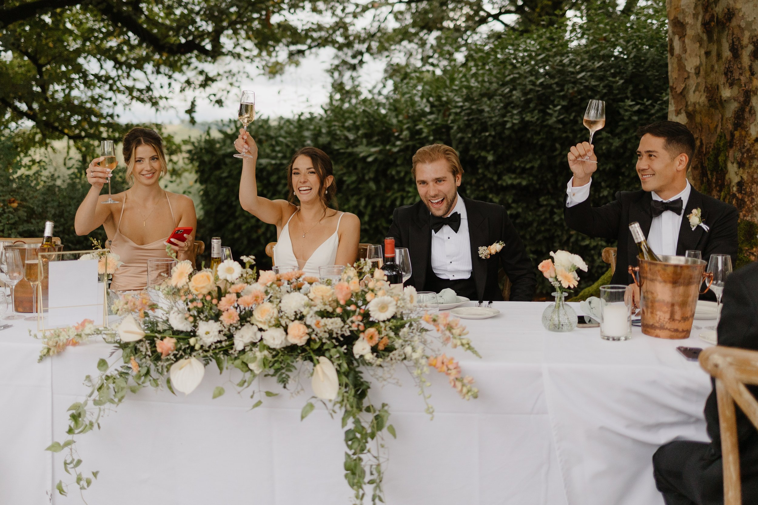 The bride and groom toast as they sit in front if their flowers of peach and white on a honor table at a wedding reception at a chateau in France for her destination wedding in France