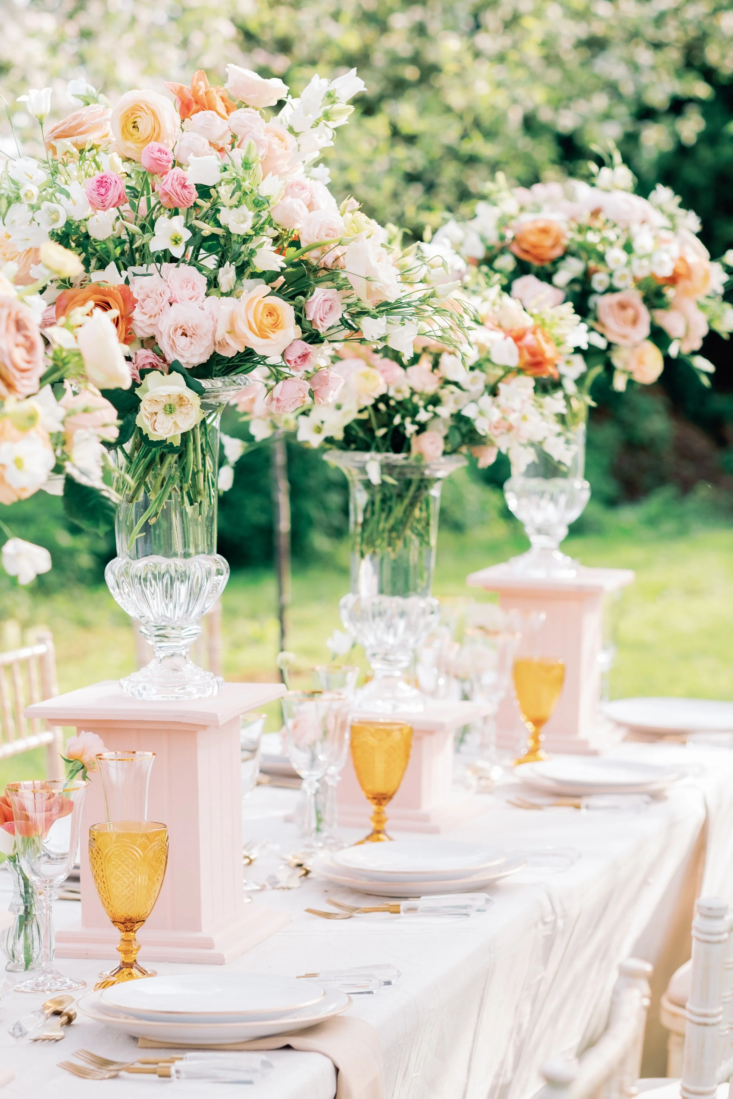 2 large wedding floral bouquets sit on a table with a pink table cloth. Flowers are springy and the background is outdoors in france