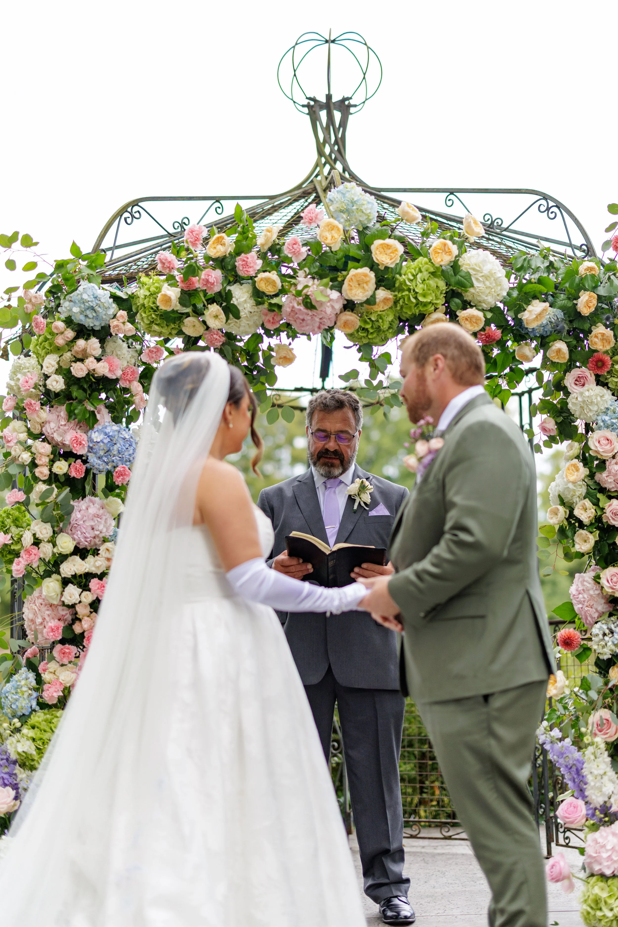 a bride and groom stand before a large floral covered arch saying their vows with a celebrant in the middle