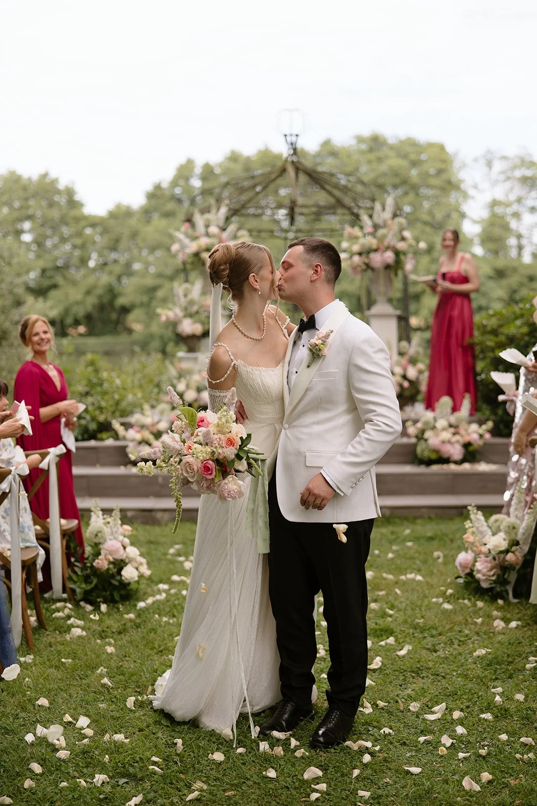 A bride and groom kiss in the ceremony aisle with a floral gazebo in the background and petals on the ground at Chateau Allure du Lac