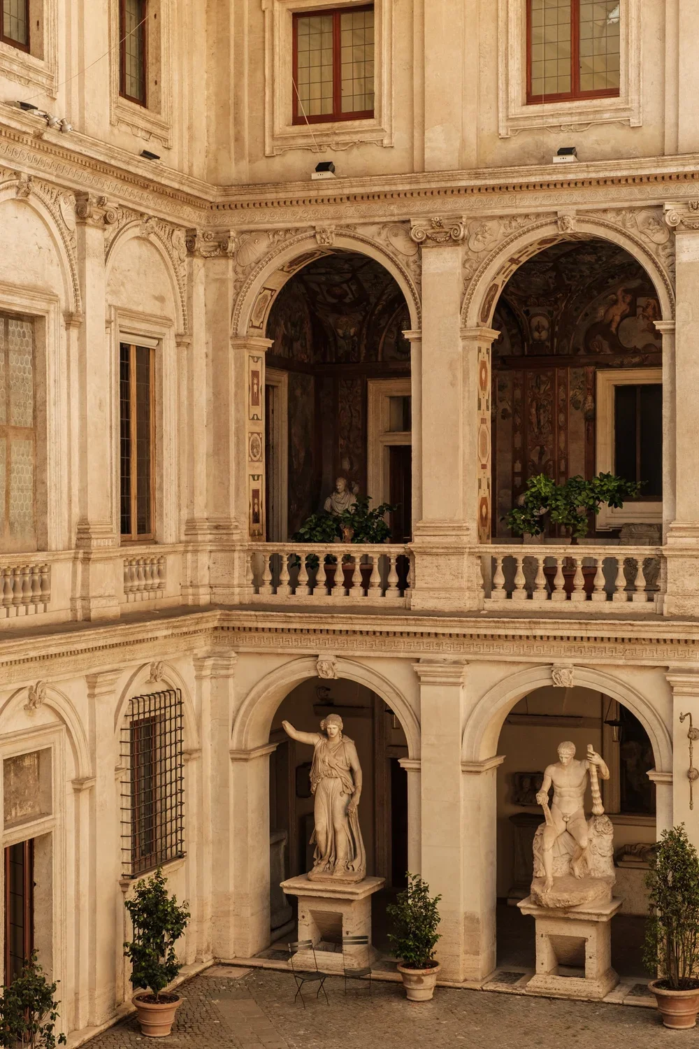 A historic European courtyard with classical architecture, statues, potted plants, and interior murals visible through open archways.