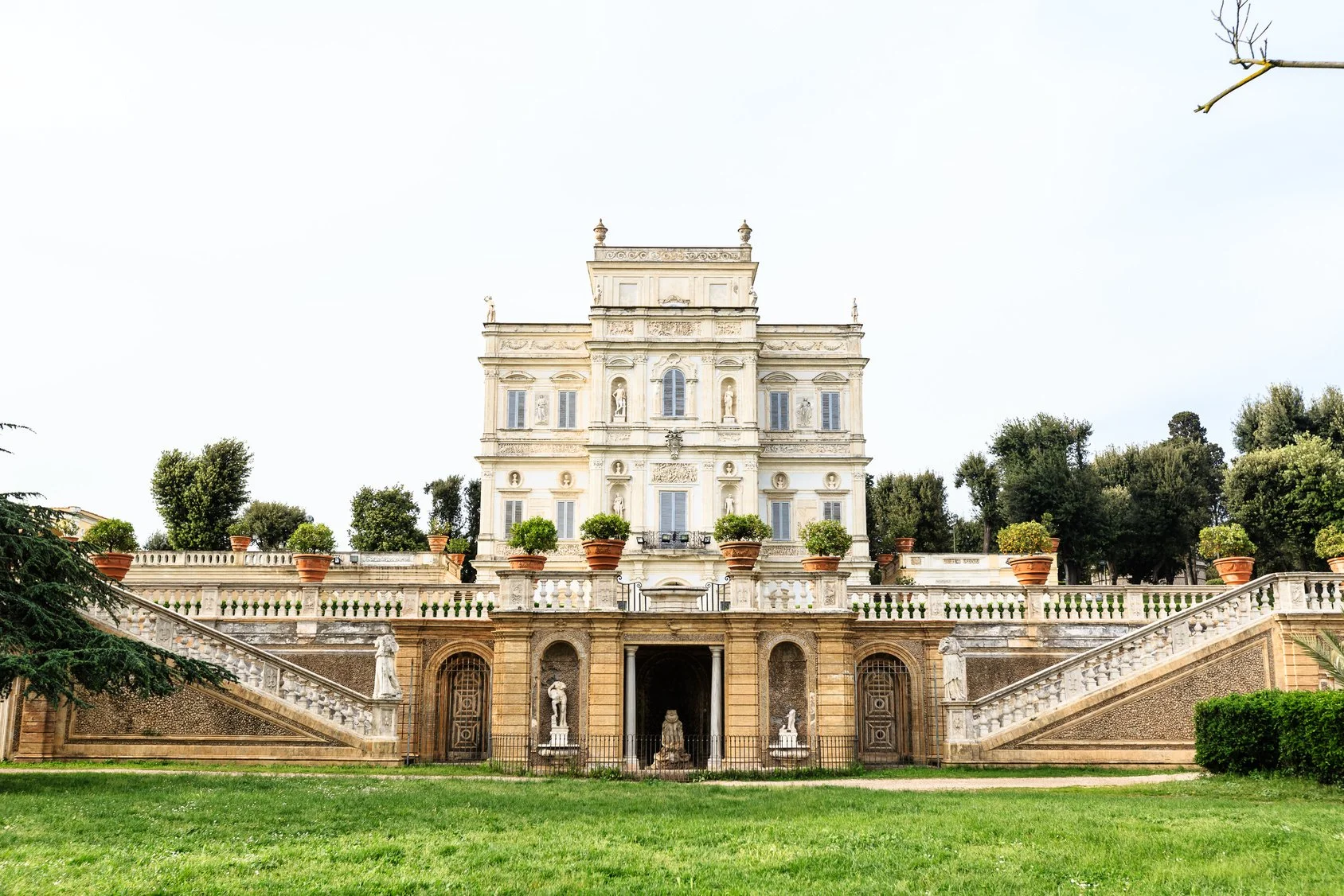 A large, ornate, white historic building with statues and plants in large pots on a terrace, surrounding staircases, and a lawn in front, with trees in the background.