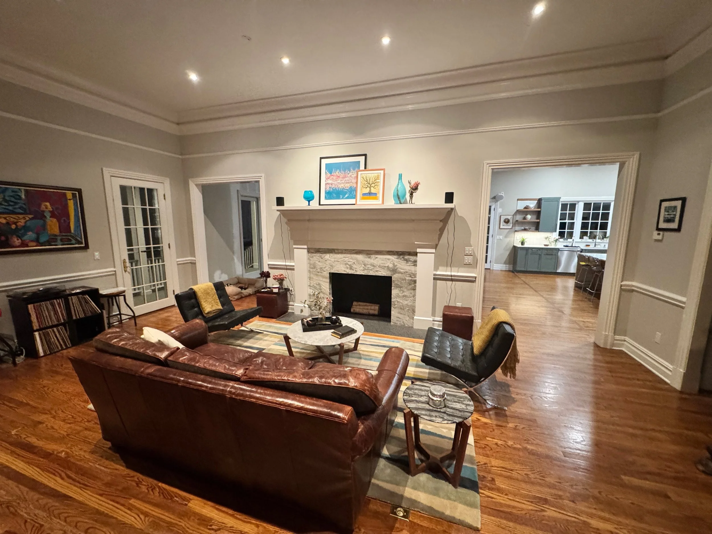 Living room with leather sofa, black chairs, fireplace, colorful artwork on wall, hardwood floors, and visible kitchen in background.