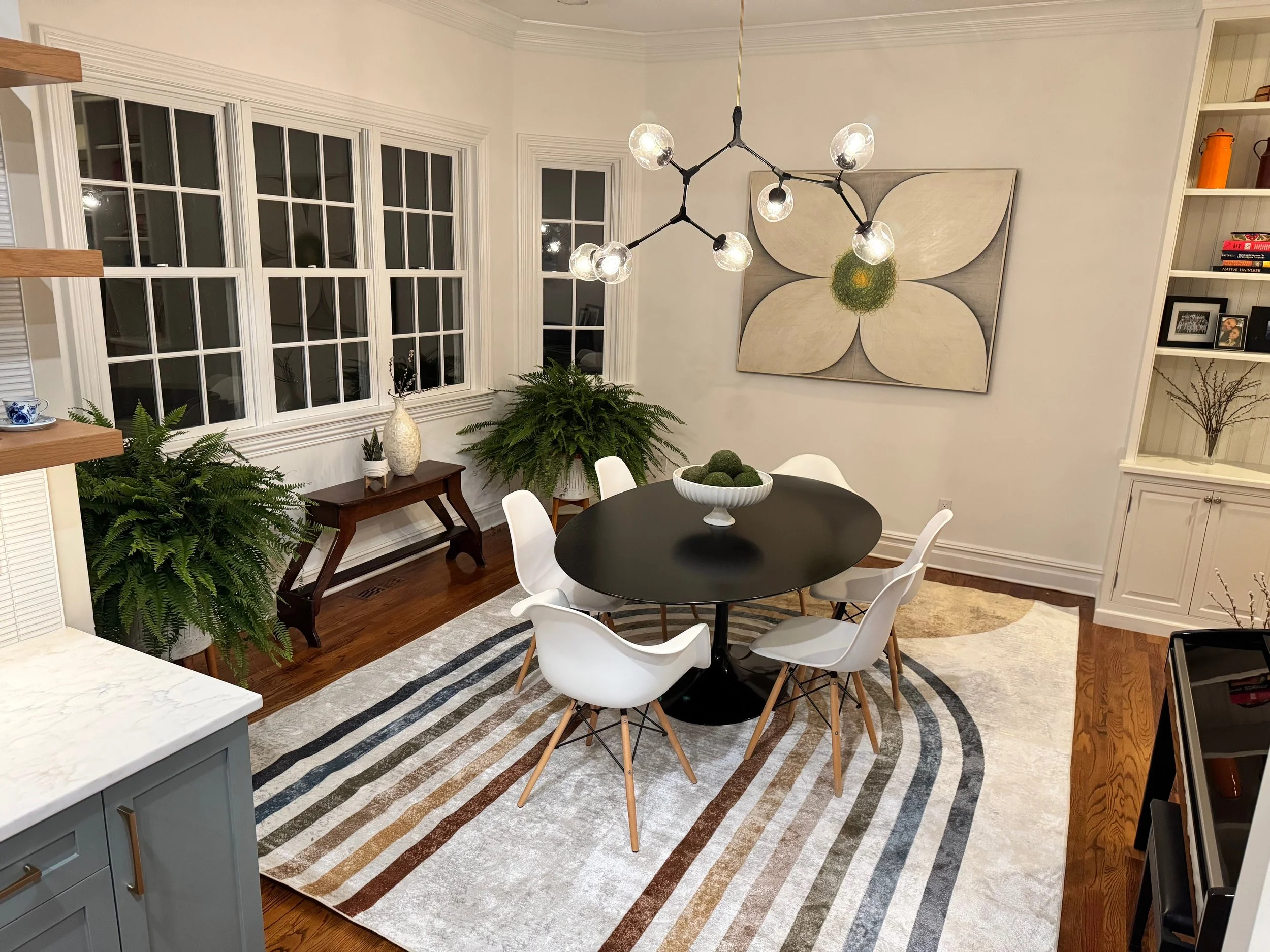 Living and dining room with black round table, white chairs, plants, large window, art on wall, chandelier, and built-in shelves.