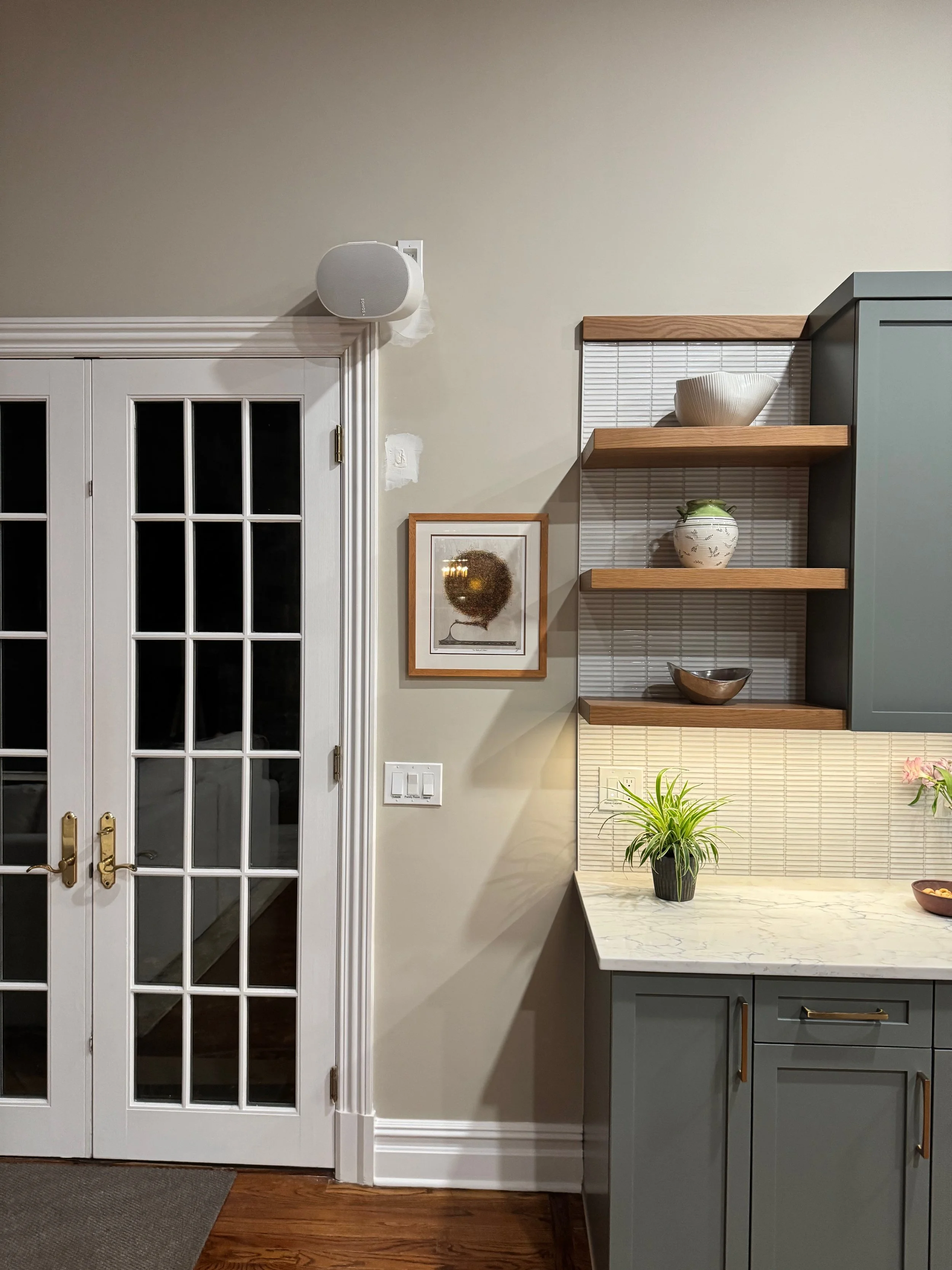 Interior scene with white double glass French doors, a framed picture of a tree, two wooden shelves with decorative bowls and a plant, a green cabinet with a marble countertop, tiled backsplash, and hardwood flooring.