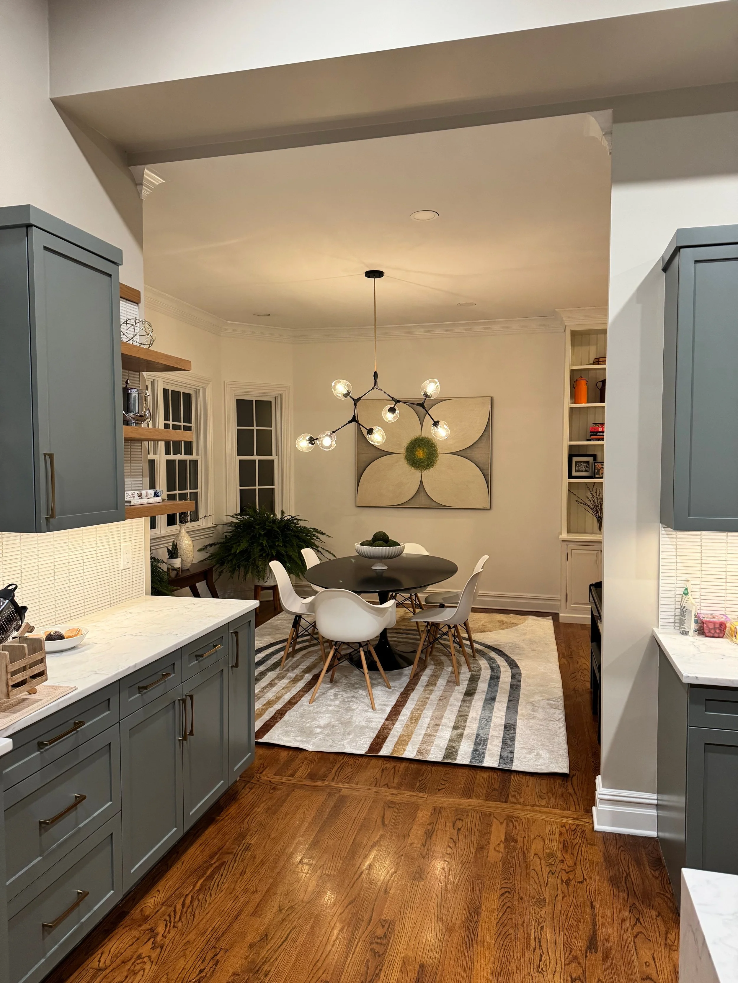 A modern dining room with a round black table surrounded by five white chairs, a geometric chandelier, a large flower artwork on the wall, a built-in bookshelf, and a plant in the corner, seen through a kitchen opening.