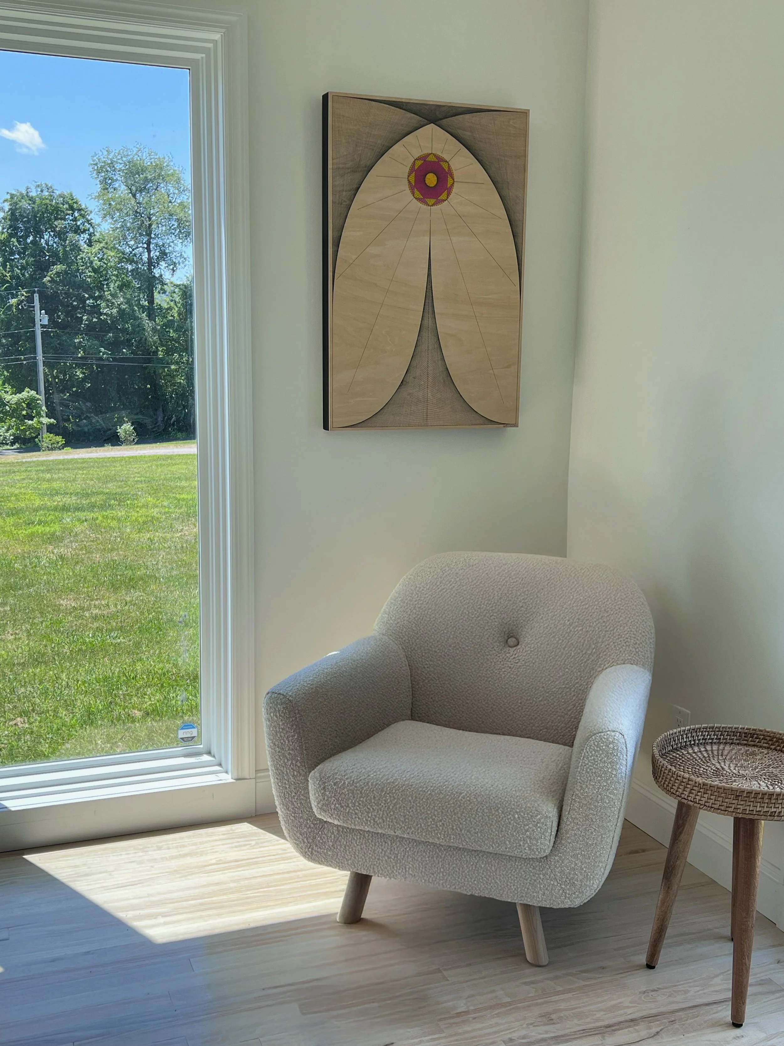 A cozy beige armchair with wooden legs next to a small wicker side table, set against a white wall with a modern abstract painting, with sunlight streaming through a large window showing a green lawn and trees outside.