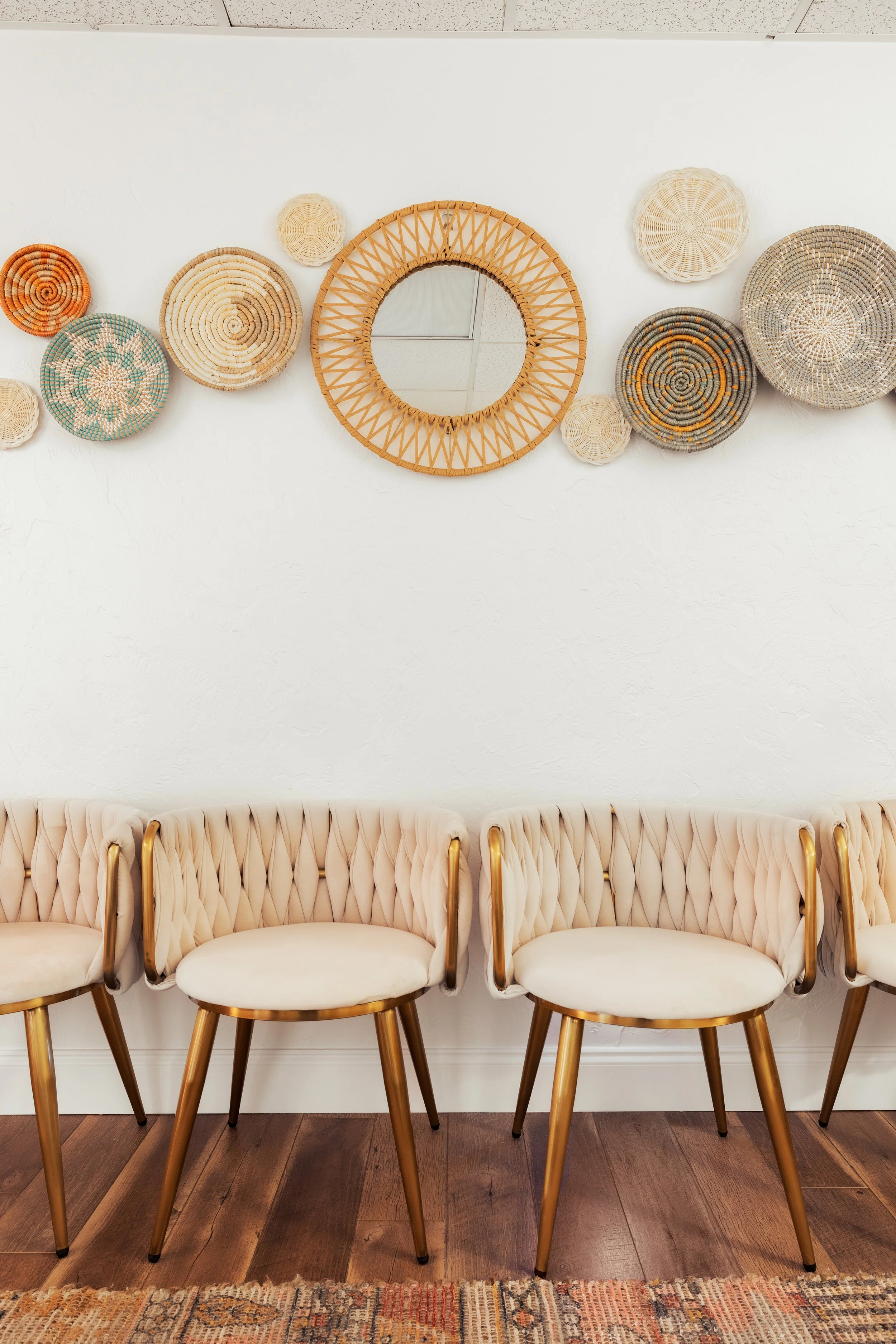Five cream-colored upholstered chairs with gold legs arranged in a row in front of a white wall. Above the chairs, there is a round mirror with a woven rattan frame surrounded by small woven baskets of various sizes and colors. The floor is dark wood