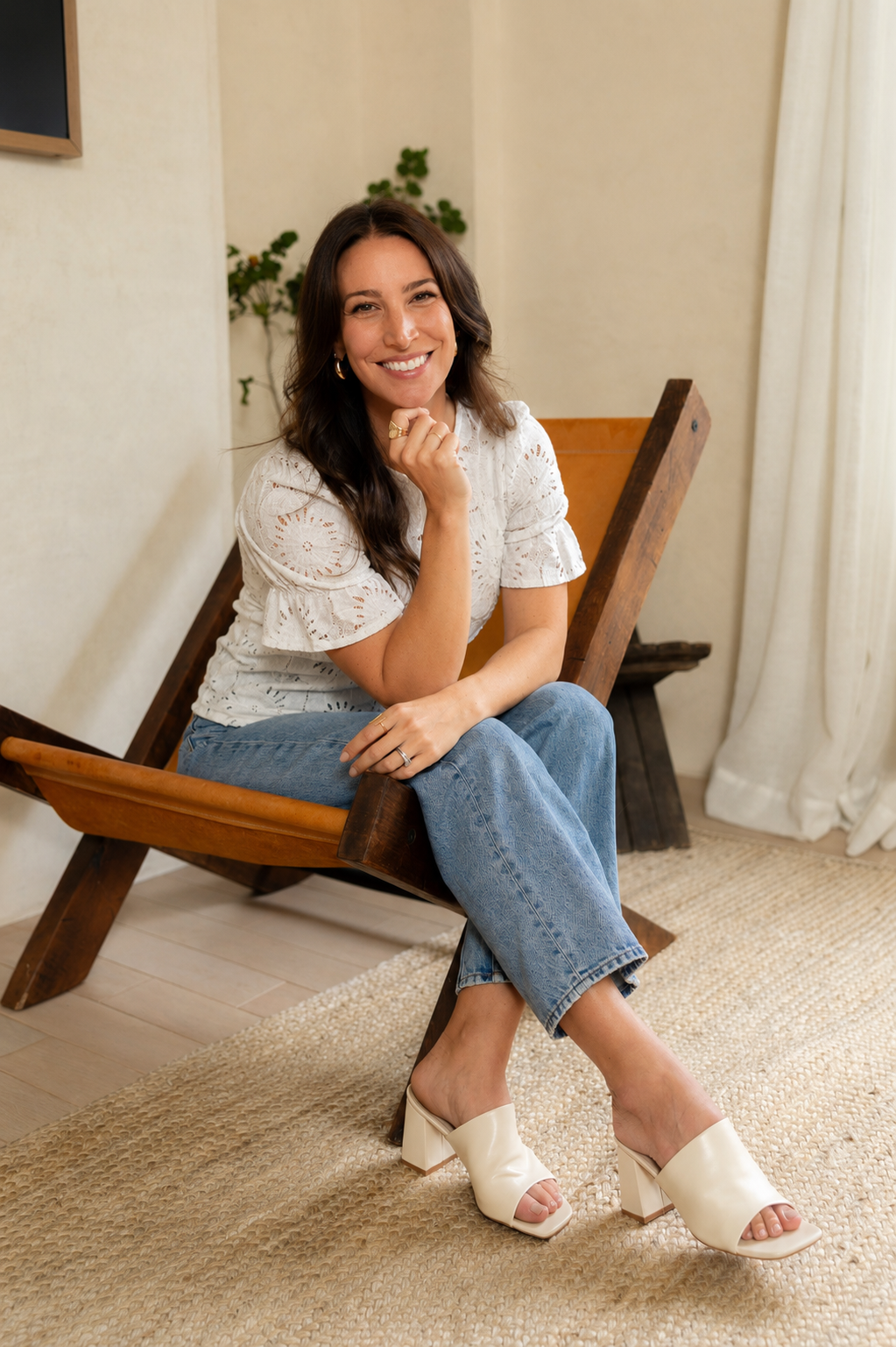 A woman sitting on a wooden bench in a cozy indoor setting, smiling, wearing a white eyelet top, blue jeans, and cream-colored high-heeled sandals.
