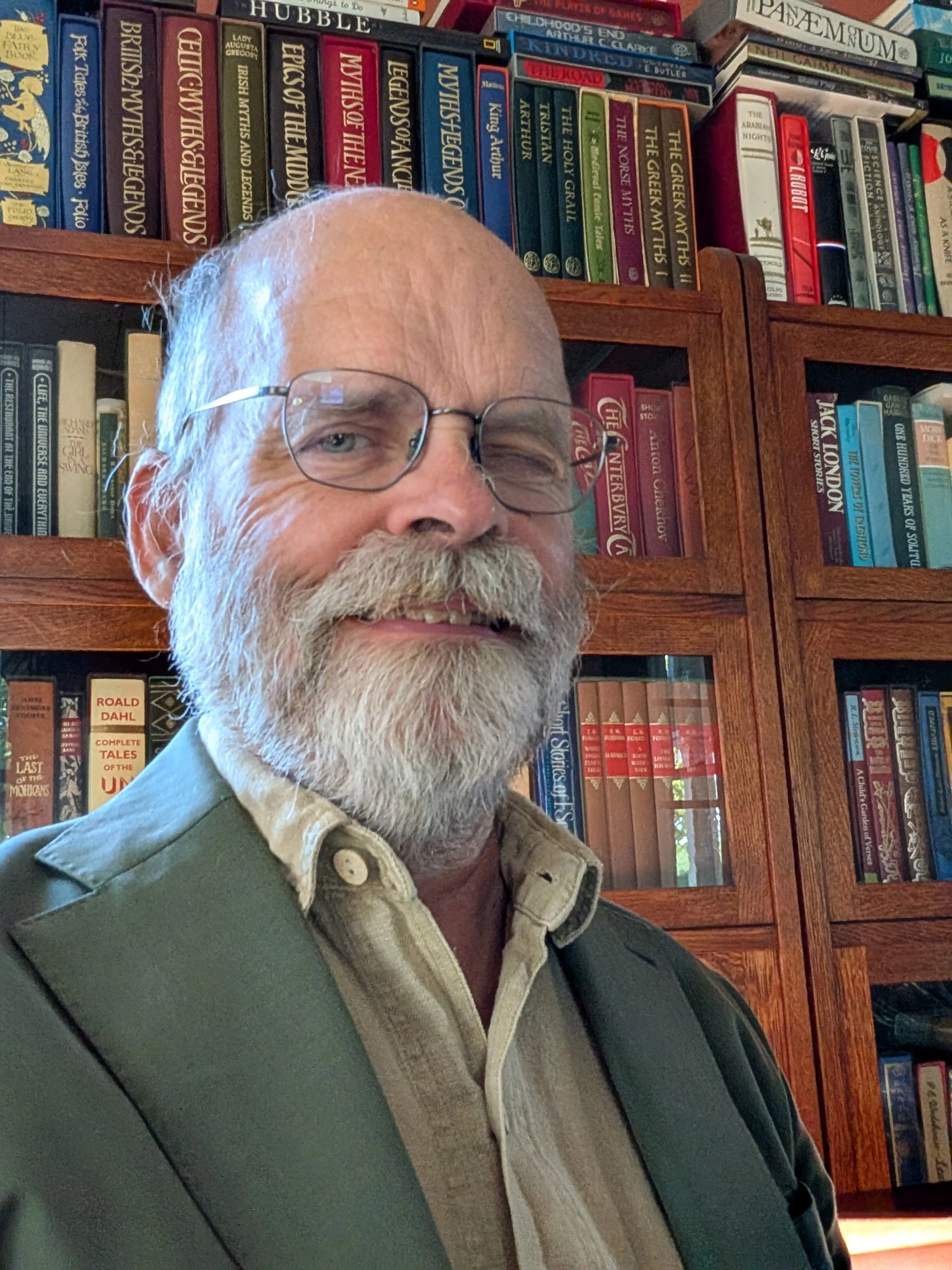 A smiling older man with glasses, white beard, and mustache wearing a green blazer over a beige collared shirt, standing in front of a wooden bookshelf filled with numerous colorful books.