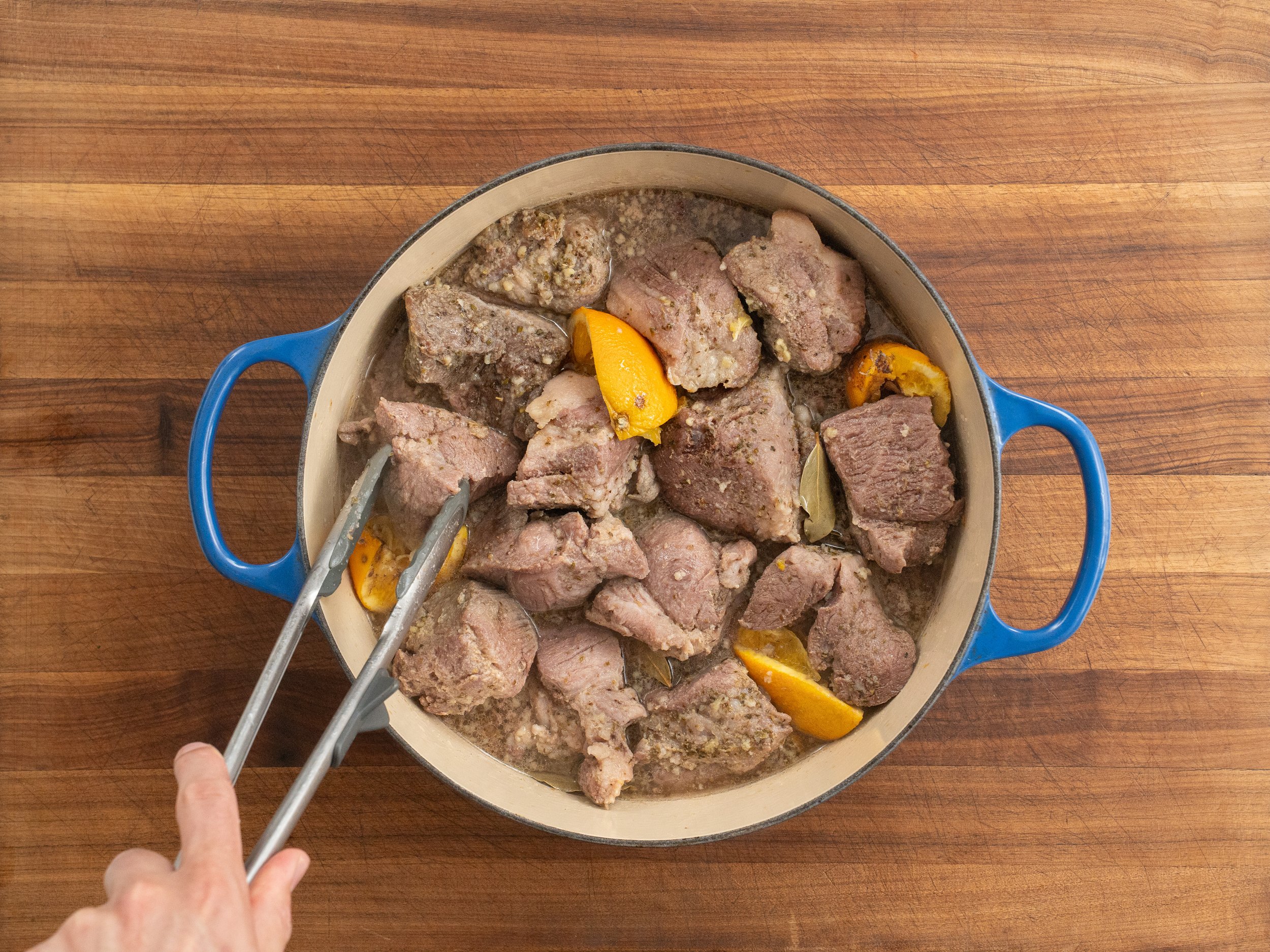 tongs flipping pieces of cooking pork shoulder in blue dutch oven
