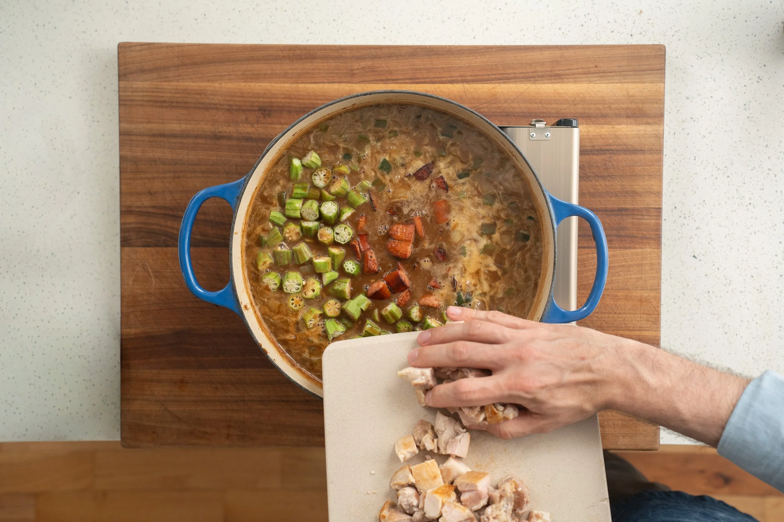 adding chicken thighs andouille and okra to a blue dutch oven filled with gumbo