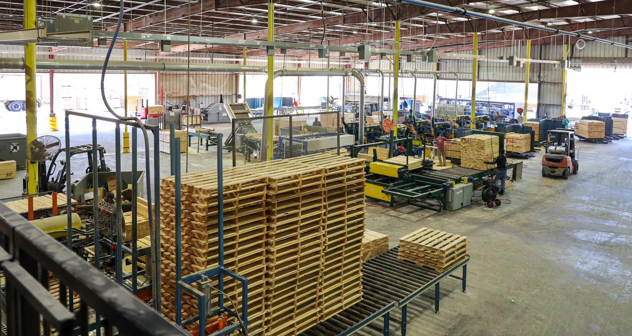 Interior of a woodworking factory with machinery, workers, and stacks of wooden pallets and boards.