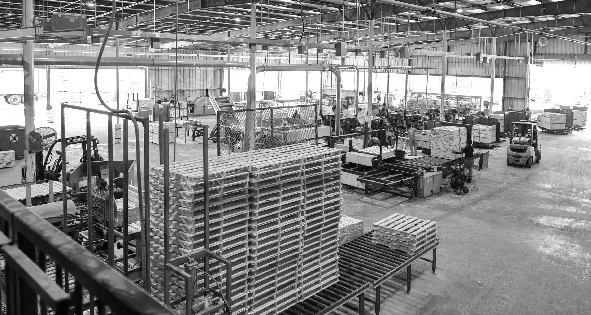 Interior of a manufacturing warehouse with machinery, workers, pallets, and forklifts in a large industrial space.