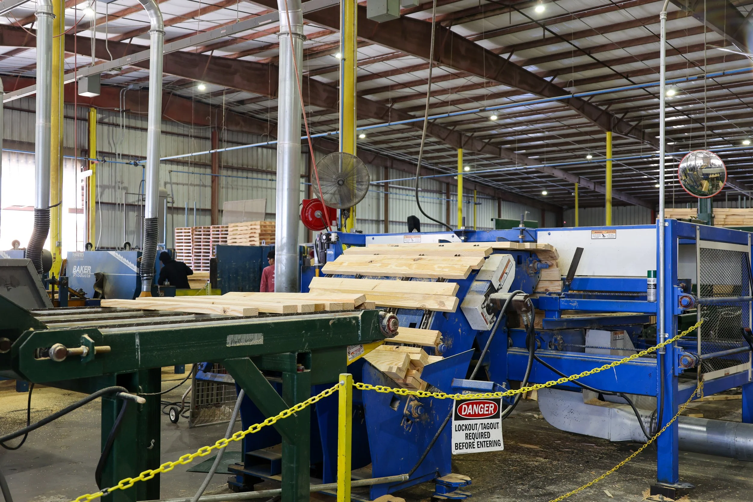Wooden planks being processed on industrial machinery inside a factory with yellow safety chains and warning signs.