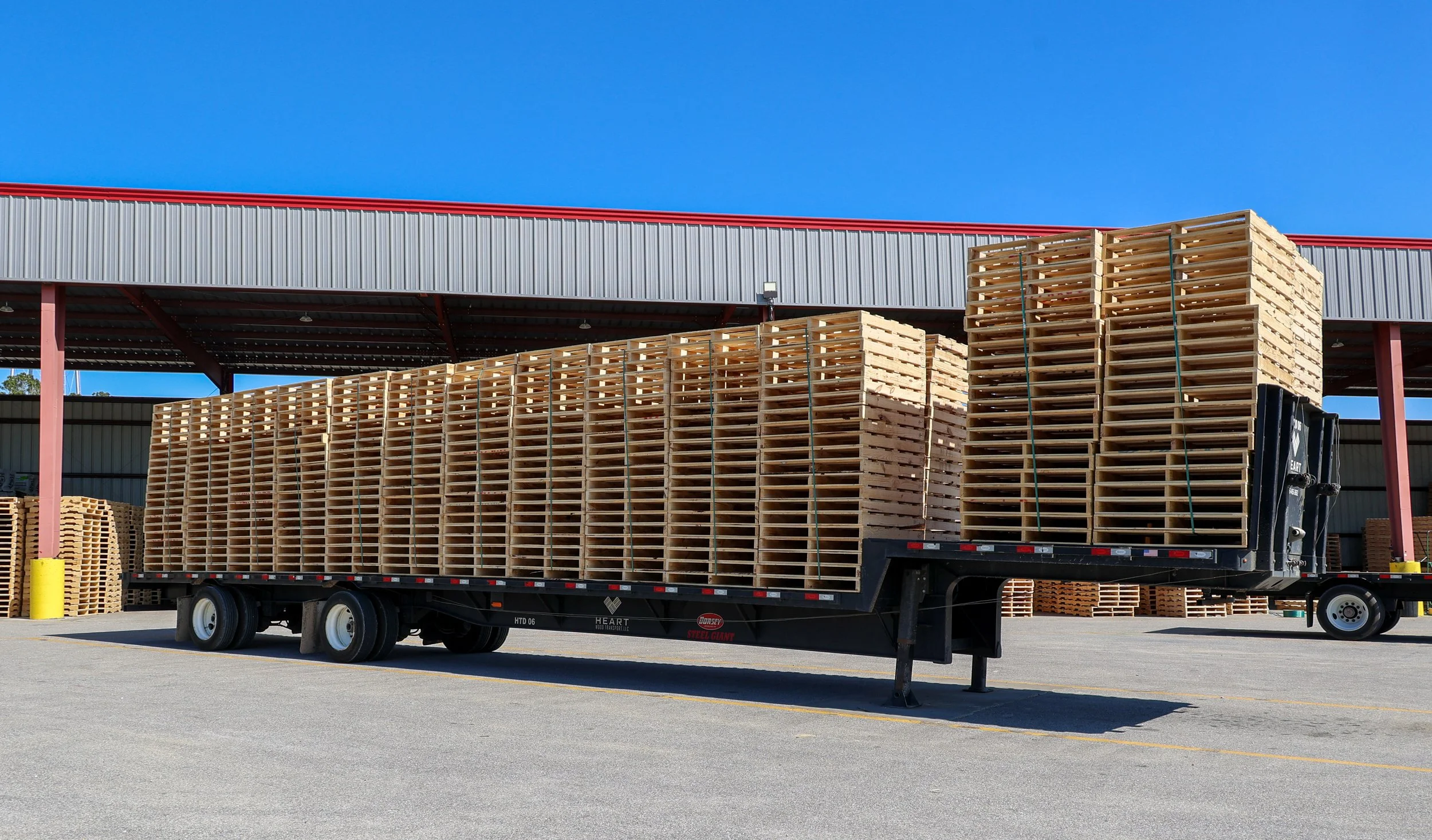 A flatbed truck loaded with stacked wooden pallets parked in an outdoor lot near an industrial building with a red roof and grey siding. Another stack of pallets is visible in the background.