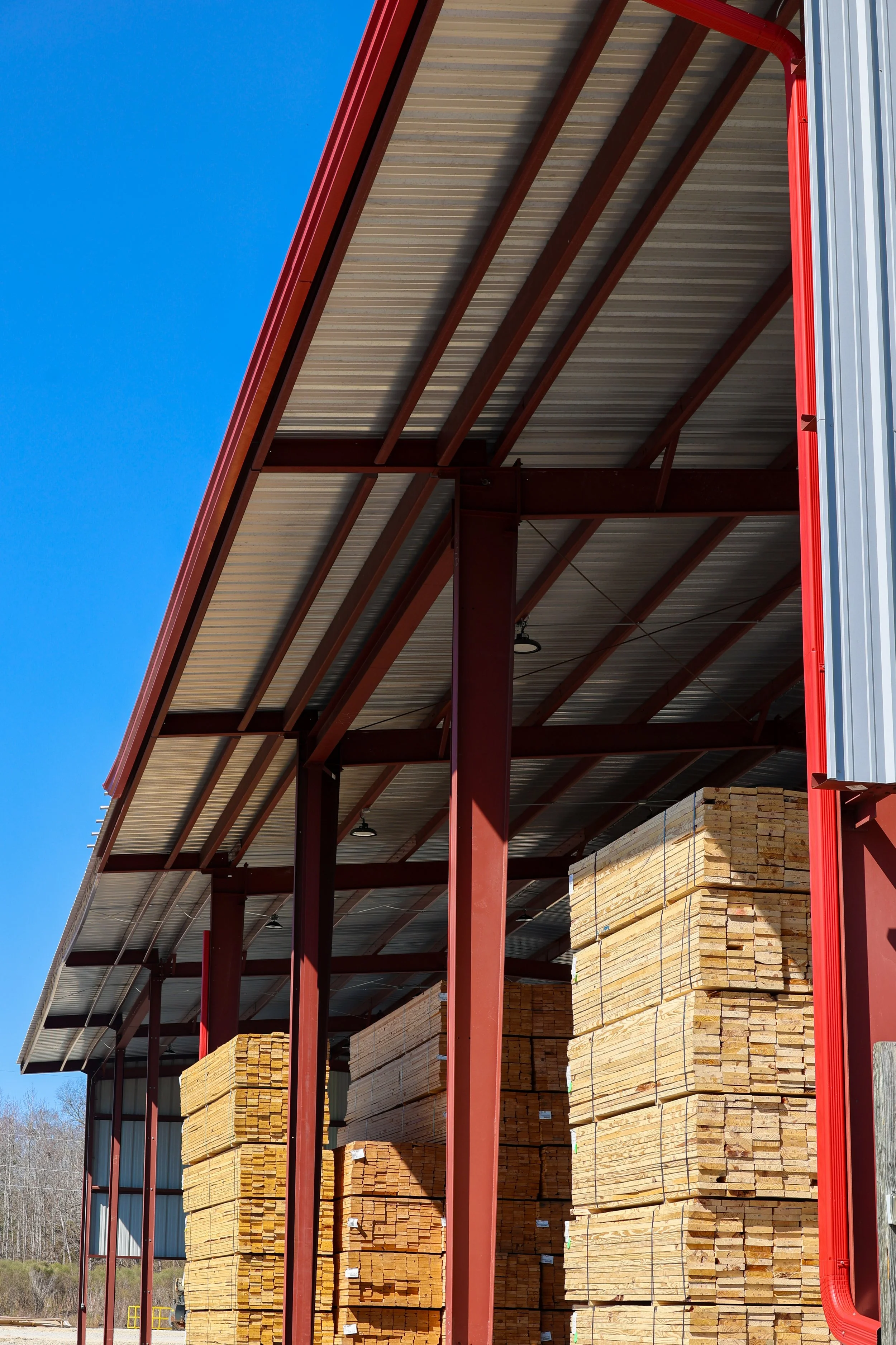 Stacked lumber stored under a red metal shed with a silver corrugated roof against a blue sky.