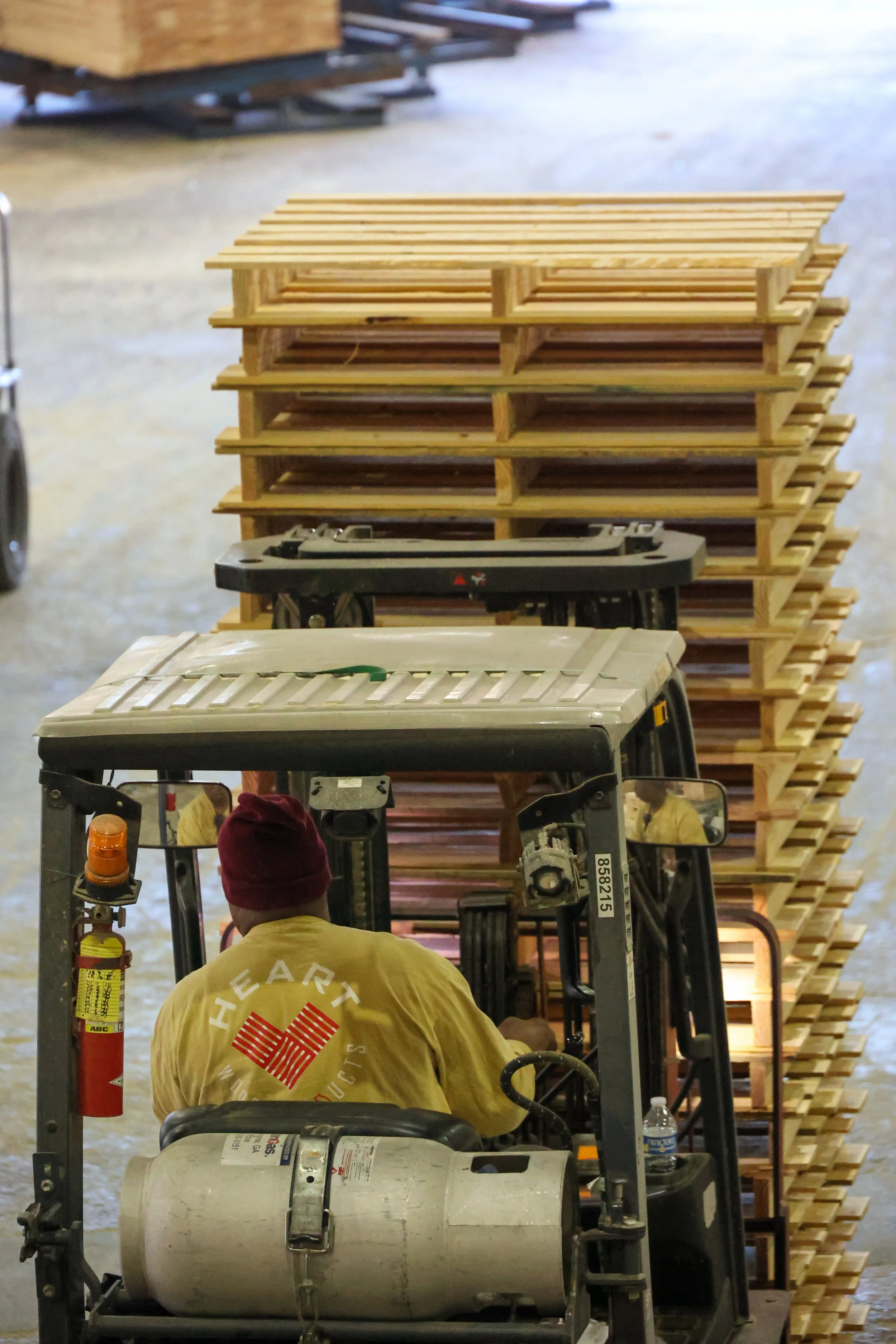 A worker driving a forklift pushes a stack of wooden pallets in a warehouse or storage facility.
