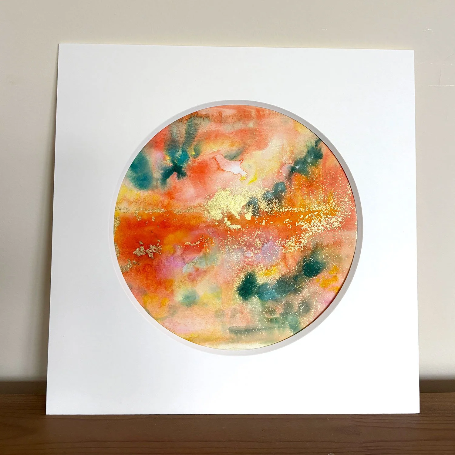 A watercolour artwork in orange, green, yellow, pink and gold. It is abstract and cosmic looking. Artwork is displayed in a soft white mount with a circular aperture