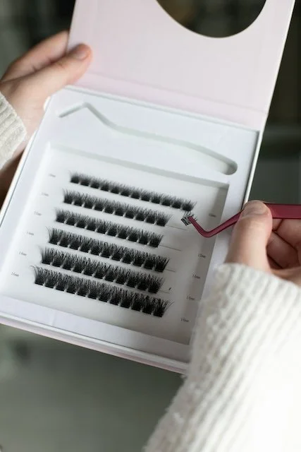 Hands holding a Lash 911 Lash Clusters kit, using tweezers to lift a small lash cluster from the tray of faux mink eyelash strips.