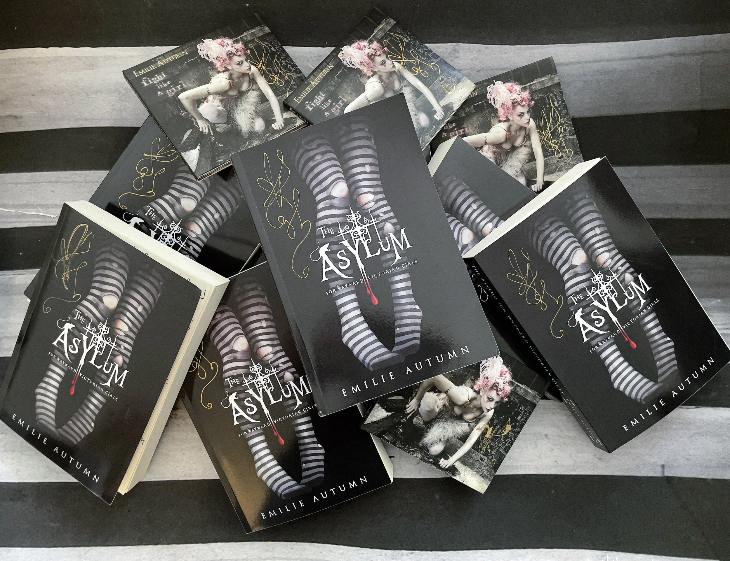Signed copies of Emilie Autumn’s “The Asylum for Wayward Victorian Girls” with autographed albums, arranged on a striped backdrop.