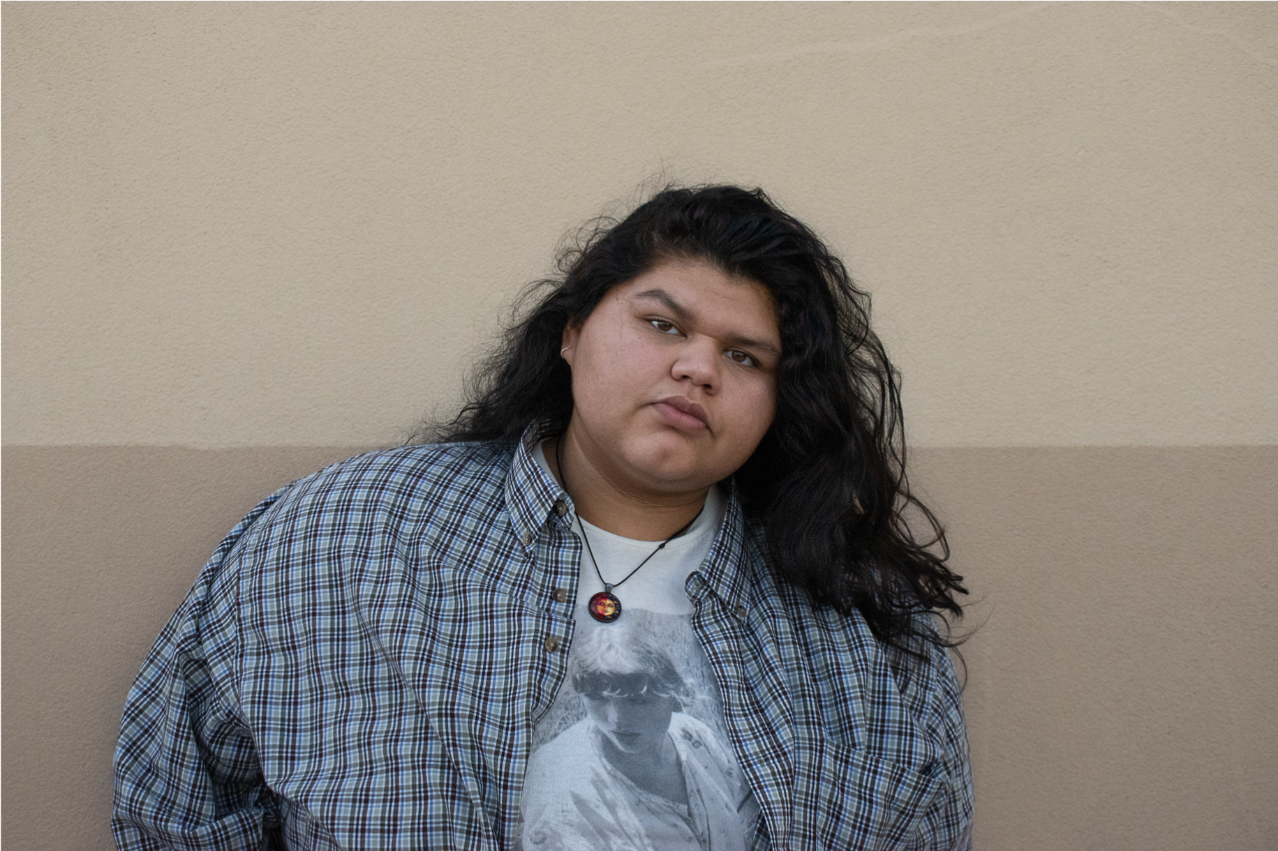 Person with long black hair, wearing a checkered shirt over a graphic t-shirt, standing against a beige wall.