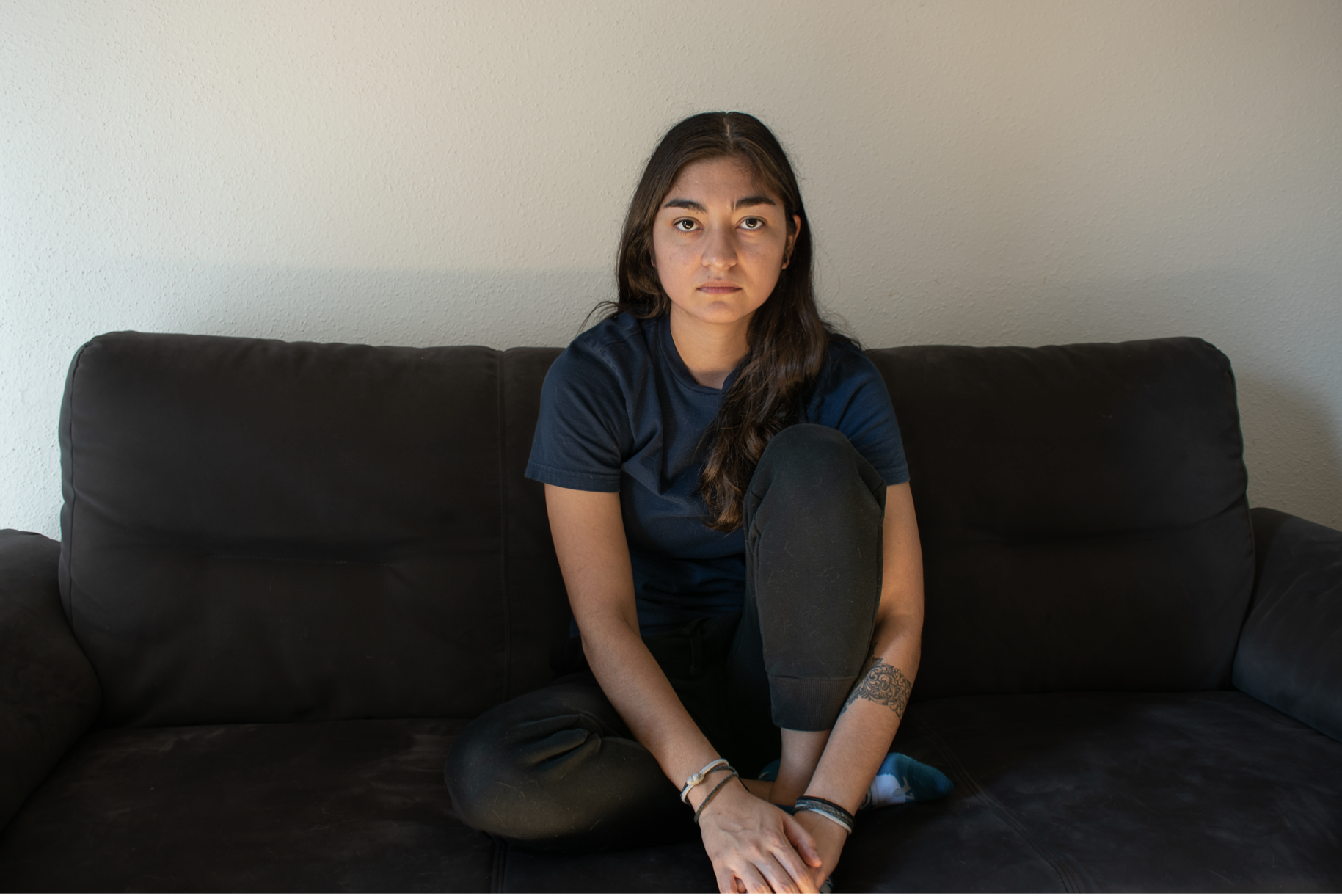 Young woman with long dark hair sitting cross-legged on a black leather sofa against a plain white wall, wearing a navy blue t-shirt and black pants.