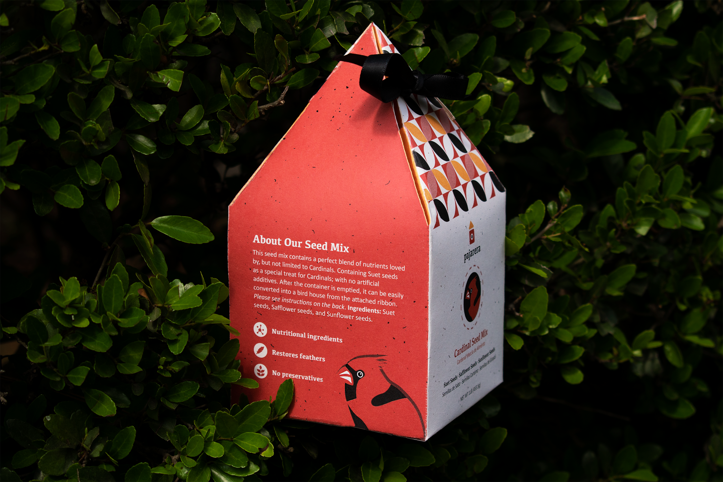 A red seed package with black ribbon attached, labeled 'About Our Seed Mix,' placed among green leaves. The package contains information about the seed mix used for planting, including ingredients and benefits.
