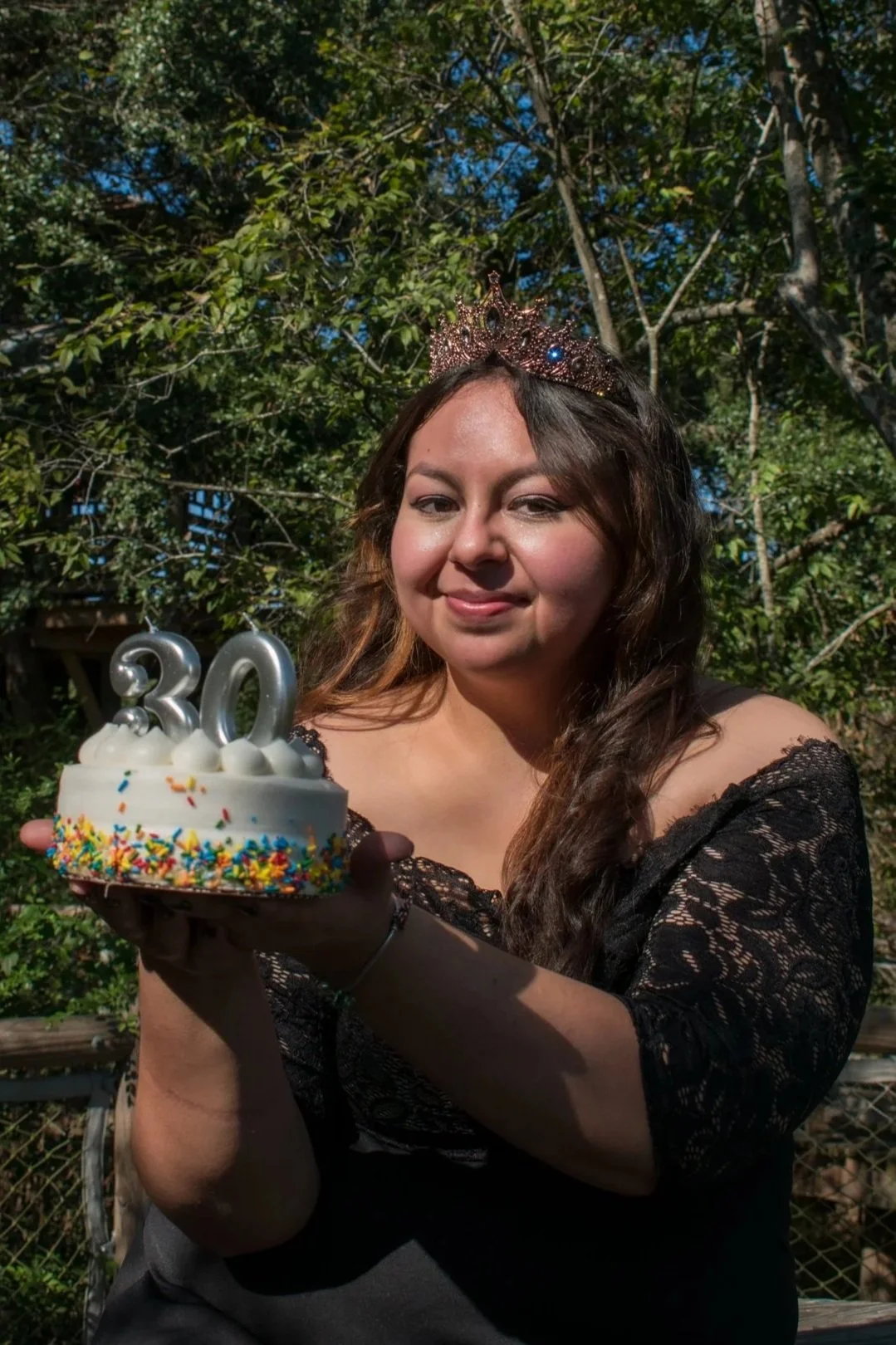 A woman with long, wavy hair wearing a tiara, holding a small birthday cake with colorful sprinkles and the number 30 candles, standing outdoors with trees in the background.