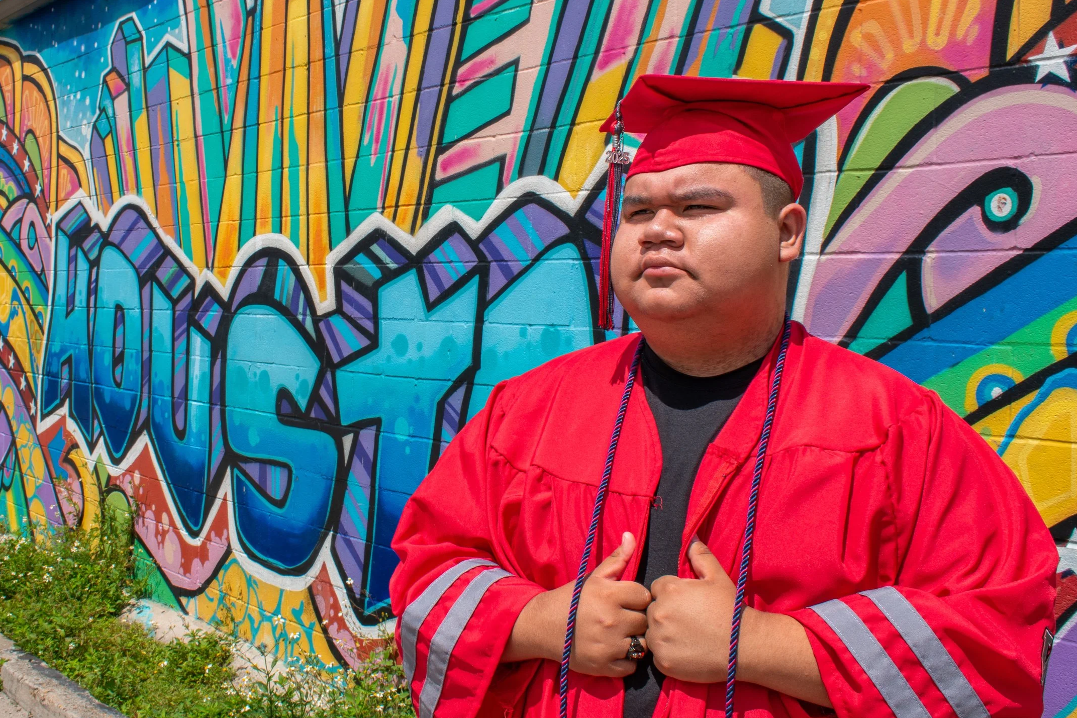 Young man in red graduation gown and cap standing in front of colorful graffiti wall, with arms crossed.