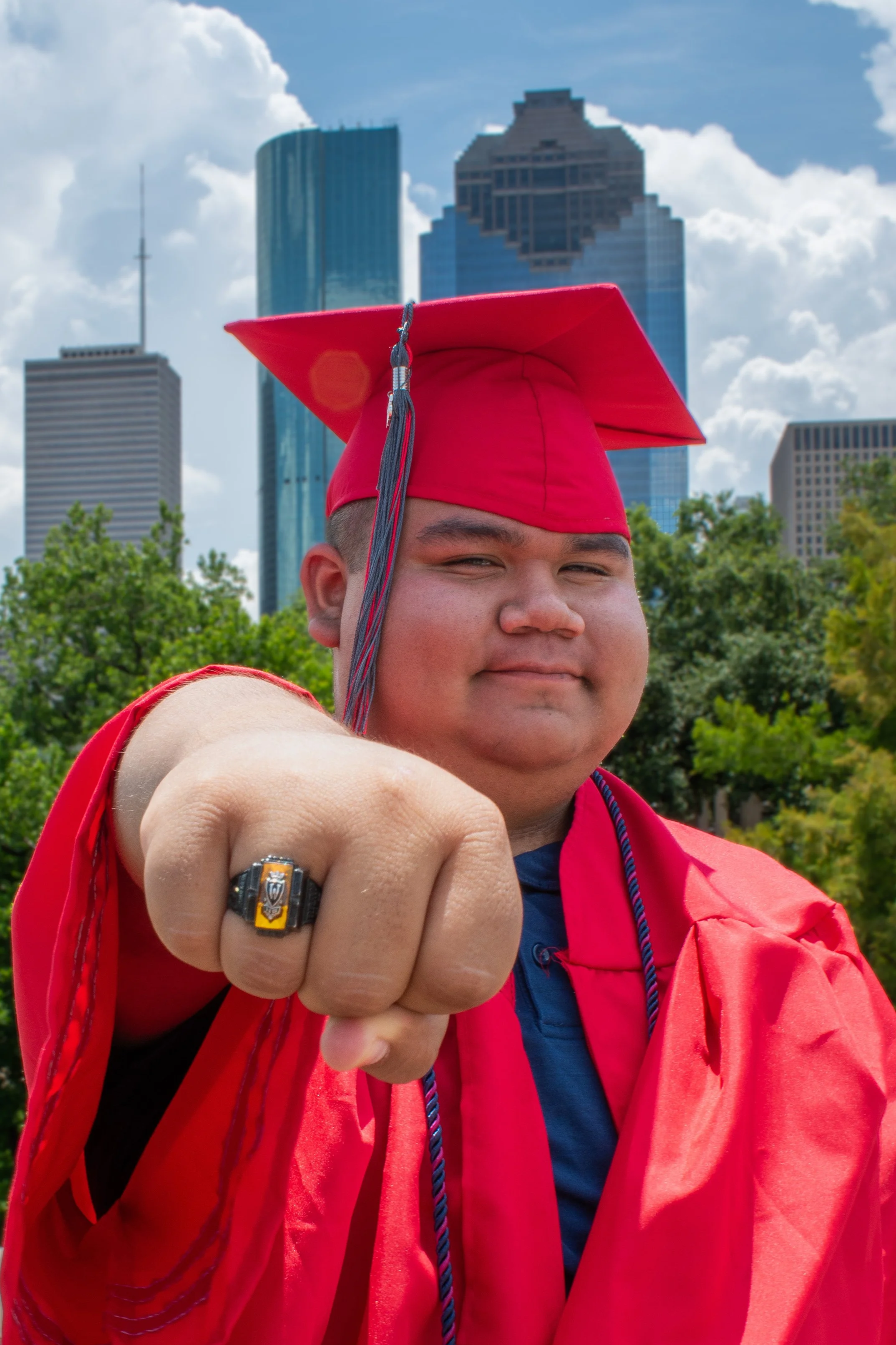 A young man wearing a red graduation cap and gown, pointing towards the camera with a confident smile. Tall skyscrapers are visible in the background under a blue sky with clouds.