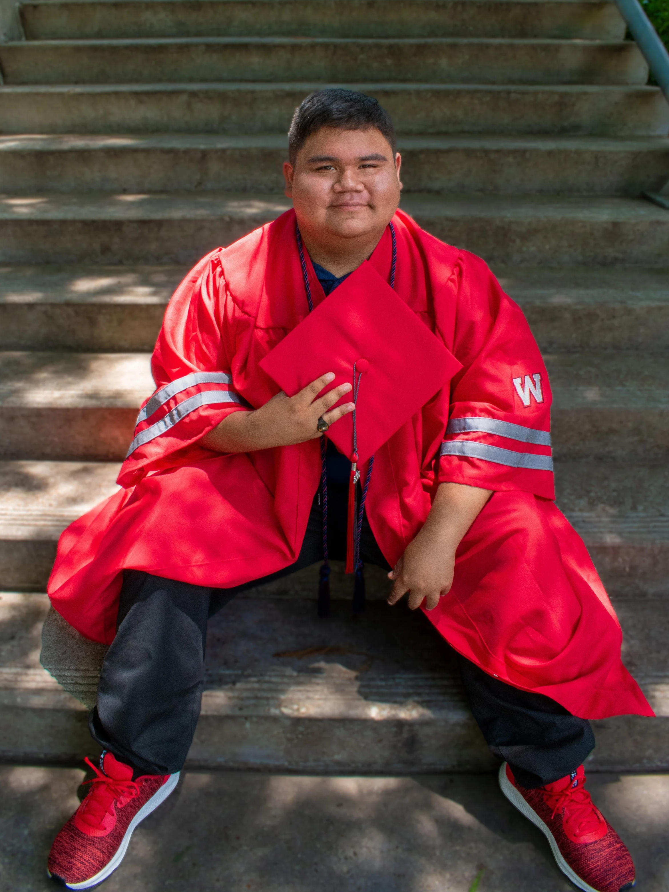 A young man in a red graduation gown and cap sitting on outdoor steps, smiling, with his hand on his chest and wearing red sneakers.