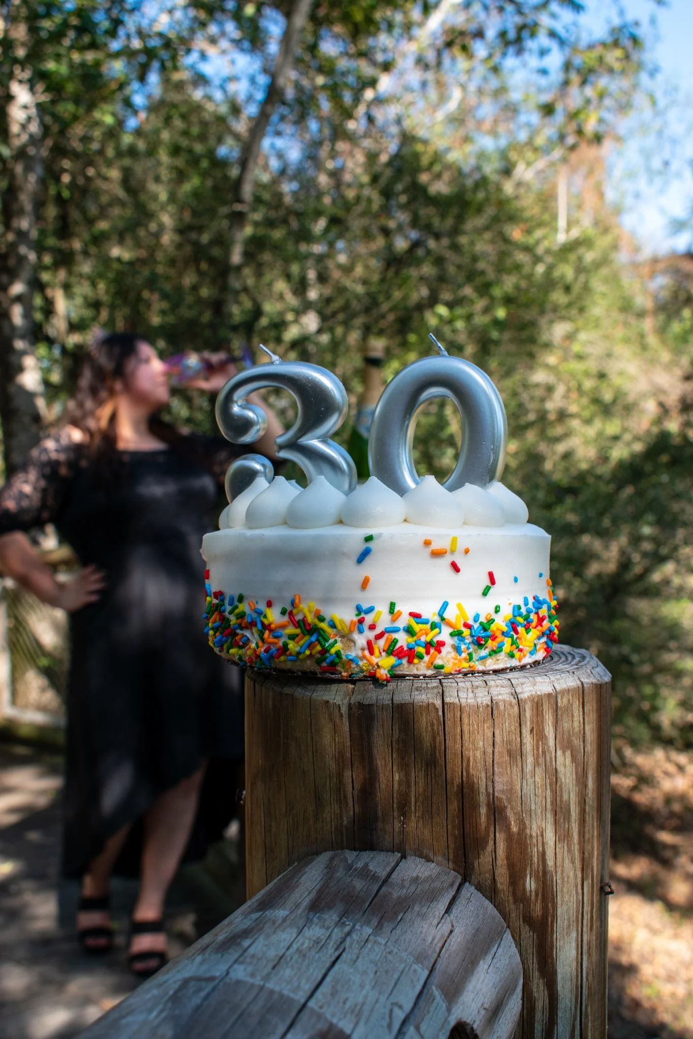 A birthday cake with colorful sprinkles and silver number candles '30', placed on a wooden post outdoors, with a woman drinking from a bottle in the background.