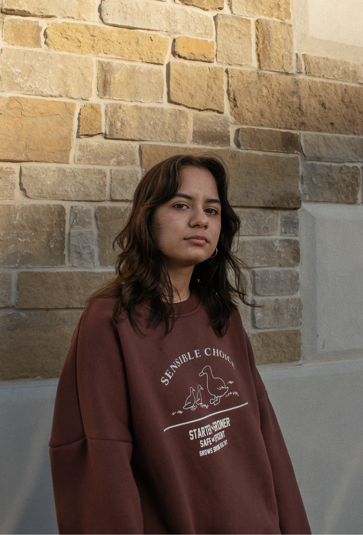 A young woman with wavy brown hair and hoop earrings stands in front of a beige brick wall. She wears a maroon sweatshirt with a graphic of ducks and the text 'Sensible Choice' on it.