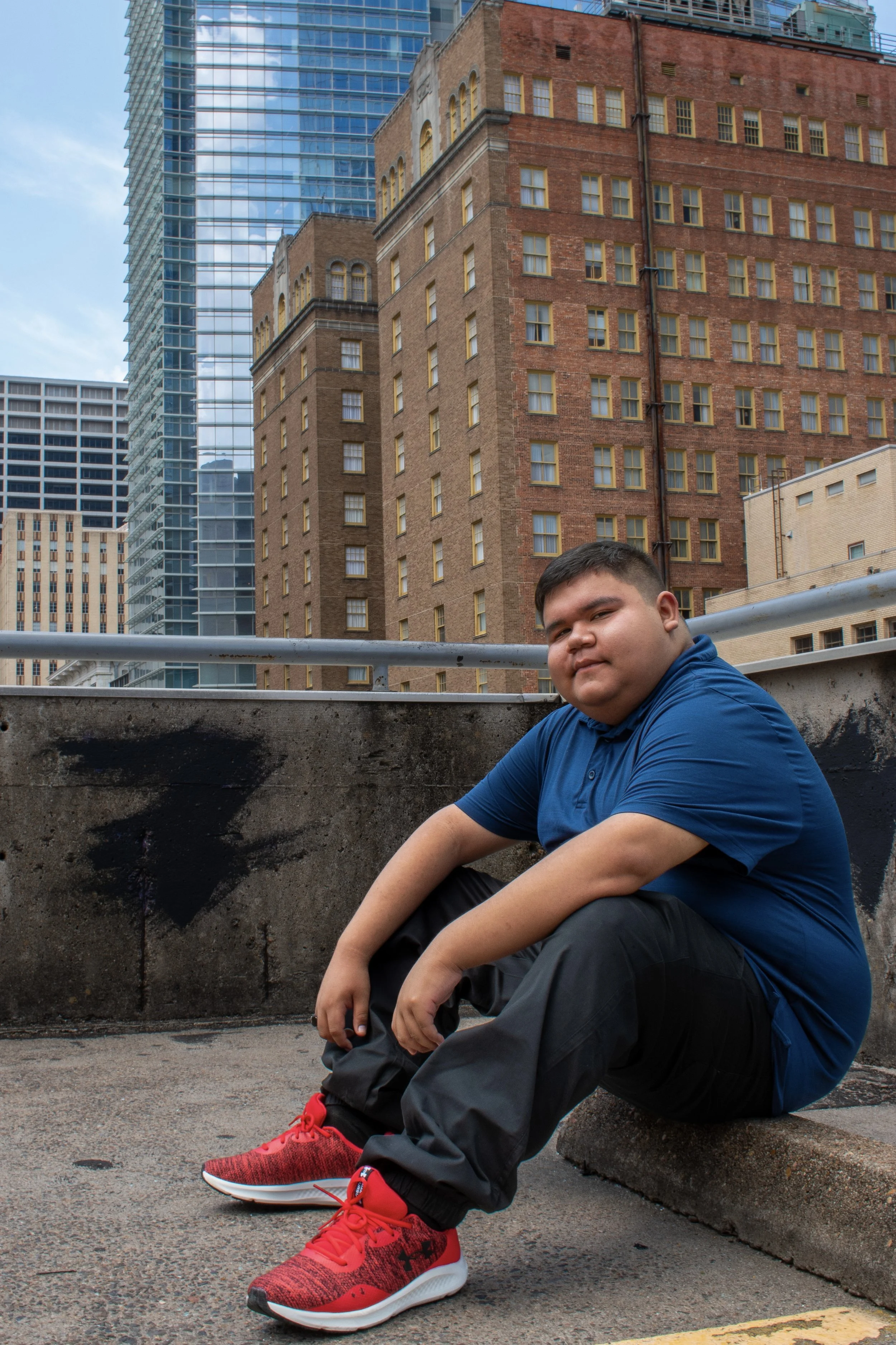 A young man with short dark hair sitting on a city sidewalk in front of a concrete barrier, wearing a bright blue polo shirt, black pants, and red athletic shoes. Tall buildings, including a red-brick building with many windows and modern glass skysc
