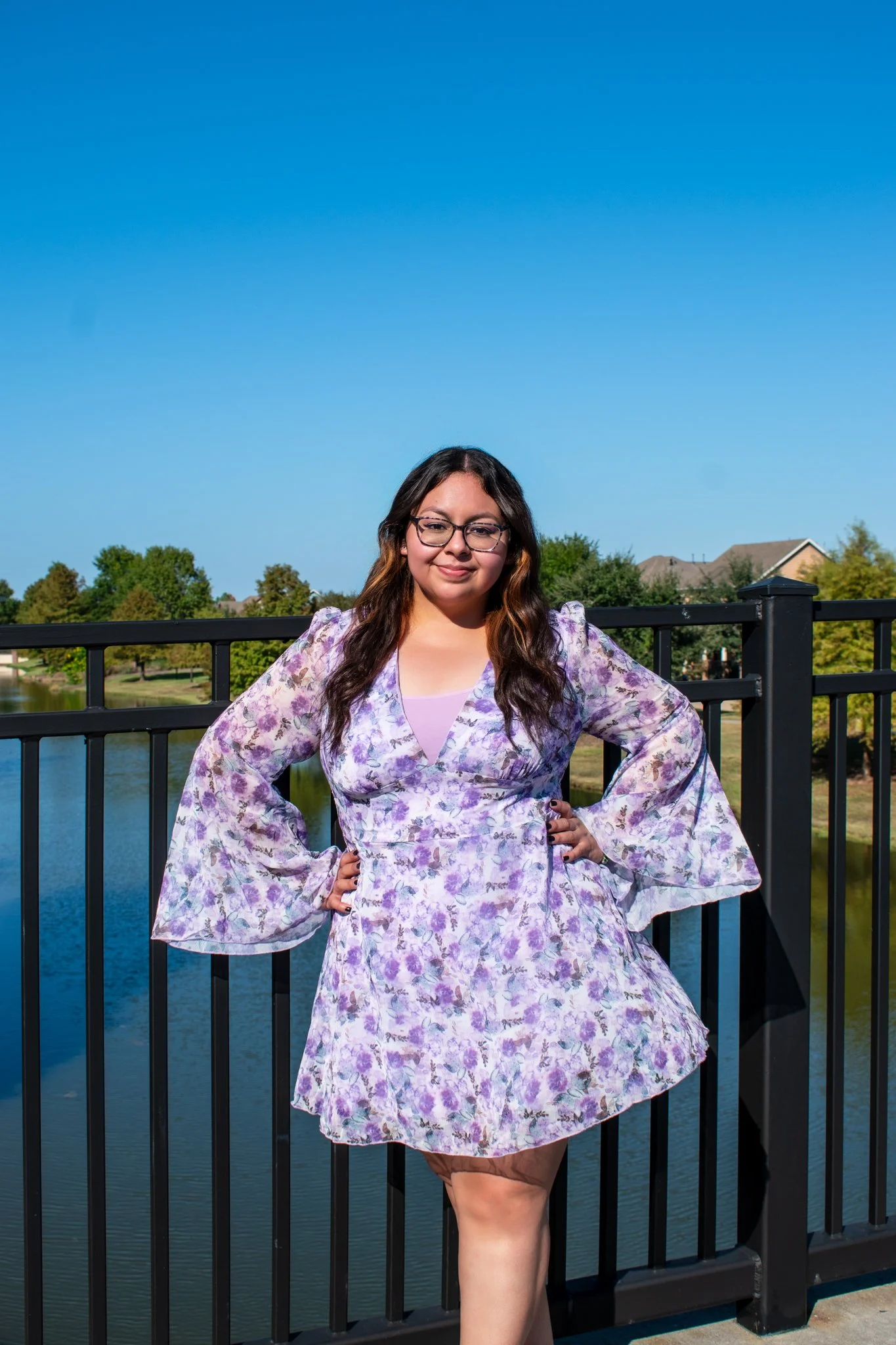A woman with long dark hair and glasses wearing a purple floral dress, standing next to a black railing with a water body and trees in the background on a sunny day.