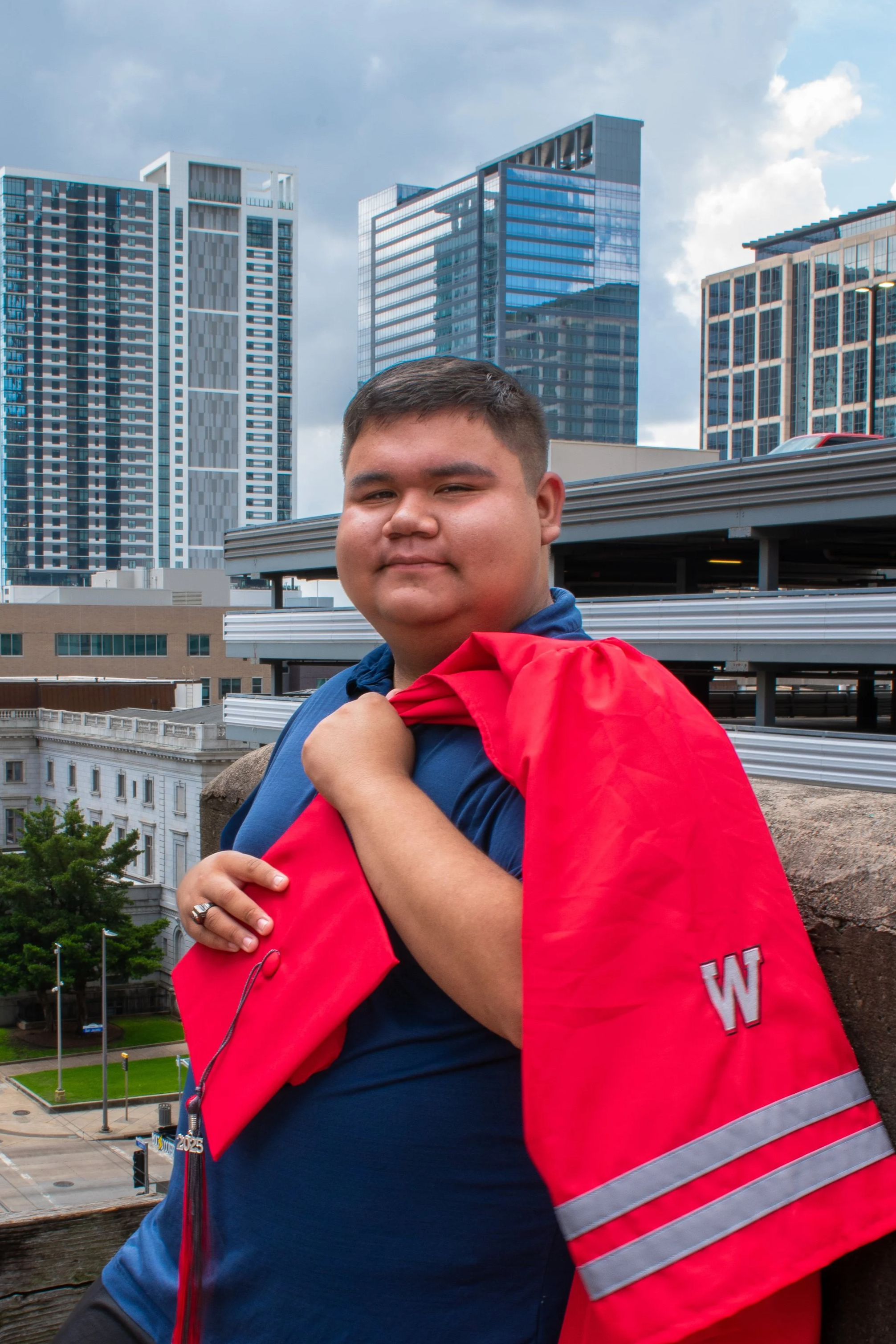 Young man holding a red jacket on a rooftop with city skyscrapers in the background.