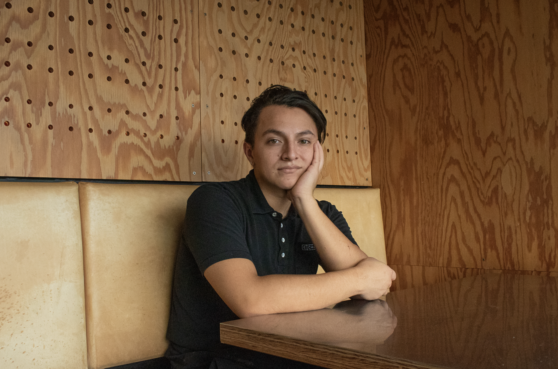 A young woman with short dark hair, resting her face on her hand, sitting at a booth in a restaurant with wood-paneled walls and beige cushioned seating.