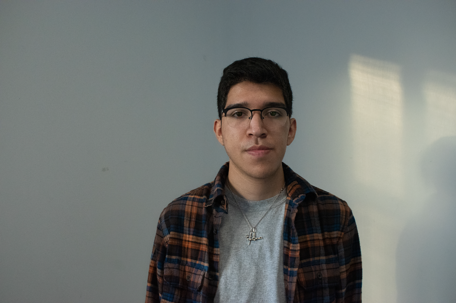 A young man with short dark hair, glasses, and light skin tone wearing a gray t-shirt, a chain necklace with a cross pendant, and a plaid shirt, standing in front of a plain light blue wall.