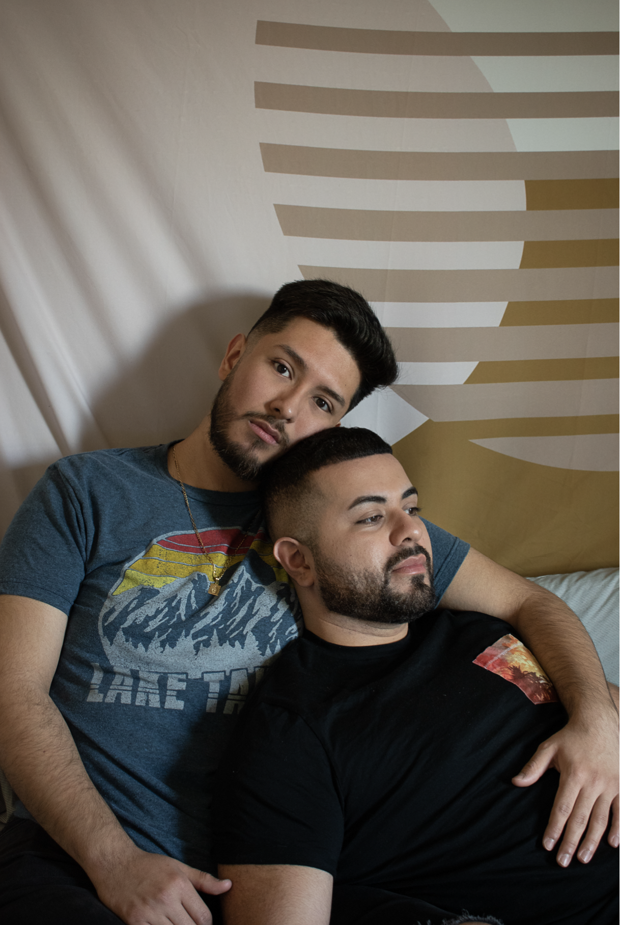 Two men sitting on a bed with a beige and brown abstract wall art in the background. The man on the left has dark hair, a beard, and is wearing a blue graphic t-shirt. The man on the right has short dark hair, a beard, and is wearing a black t-shirt.