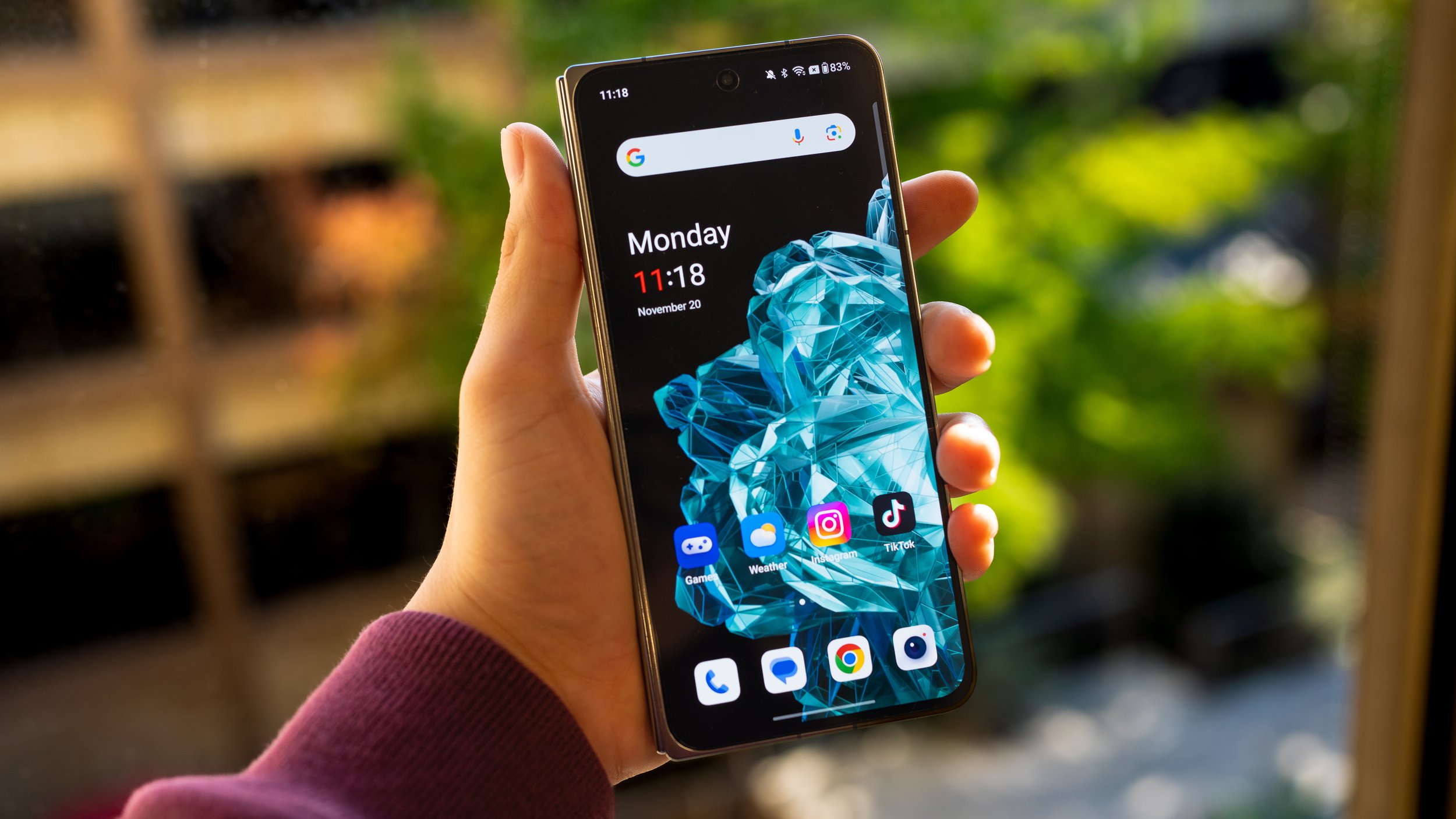A person holding a smartphone outdoors, showing a home screen with the time 11:18, date November 20, and various app icons on a background of a blue geometric design.