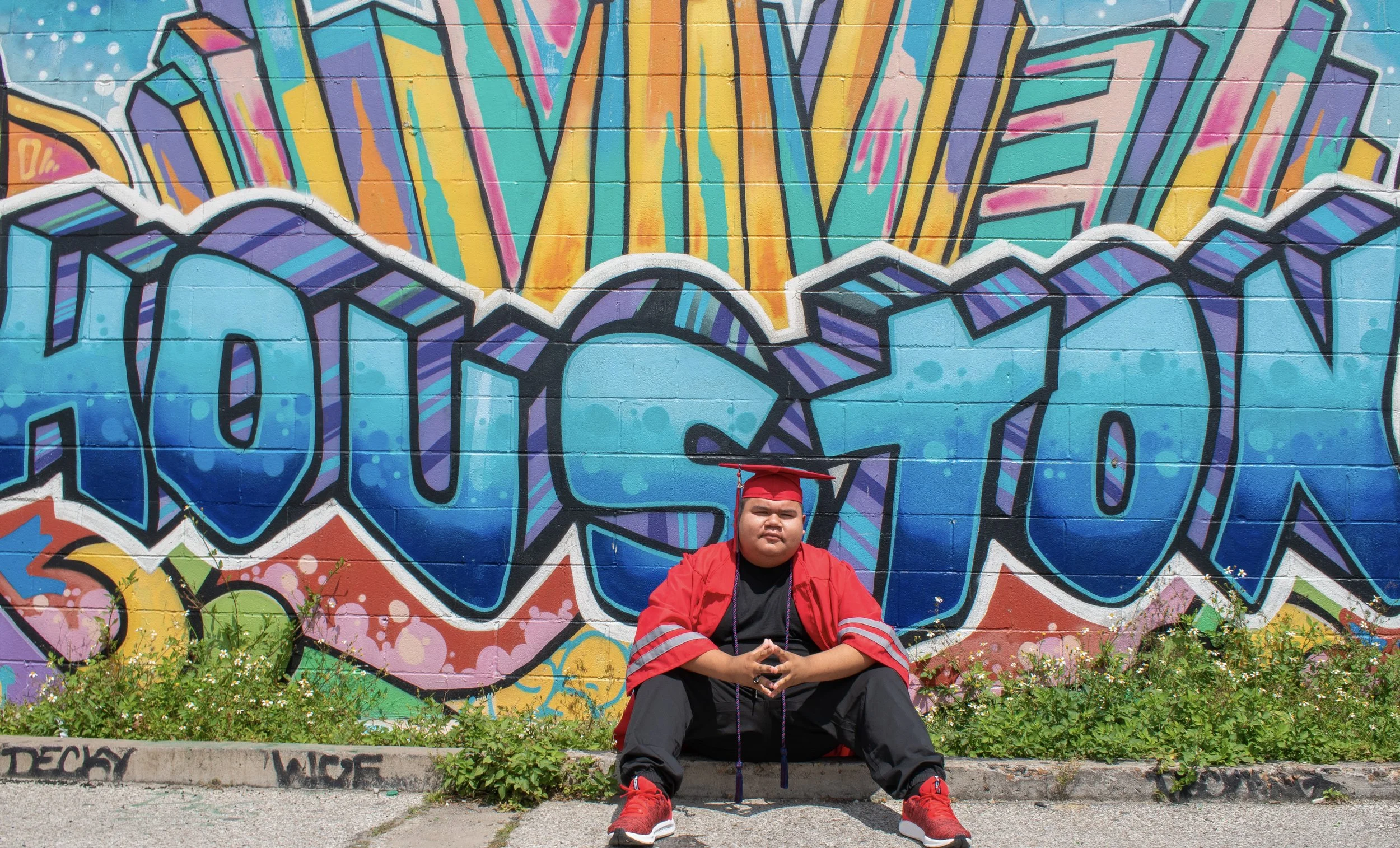 A person sitting on the sidewalk in front of a colorful graffiti wall, wearing a red jacket, black pants, and red sneakers, with a black graduation cap.